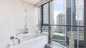 Un baño moderno con una bañera grande, una pared de mármol y vistas a edificios altos a través de amplias ventanas.