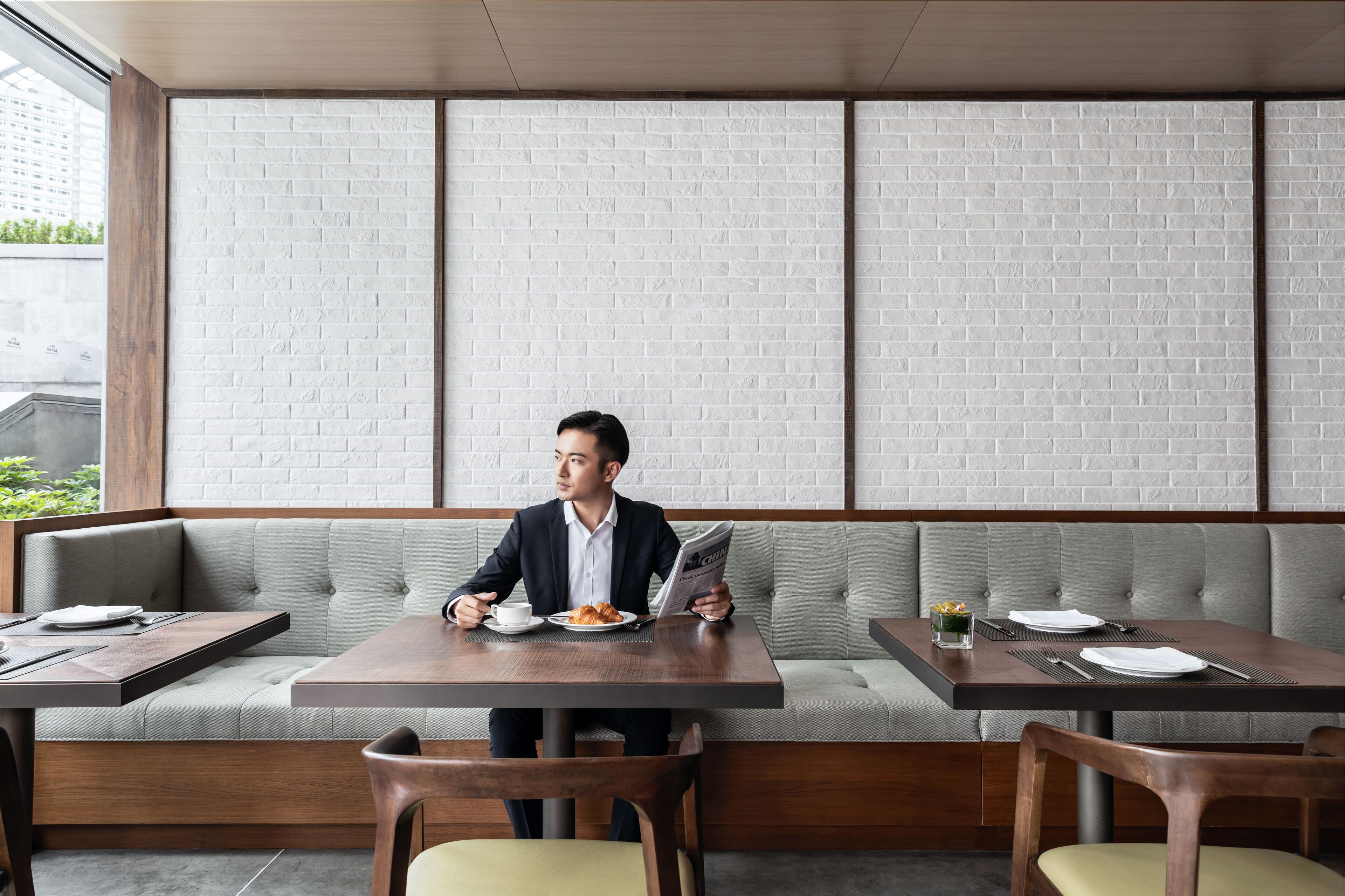 Een man zit aan een tafel in een restaurant, met een bakstenen muur, een bank en een raam op de achtergrond.