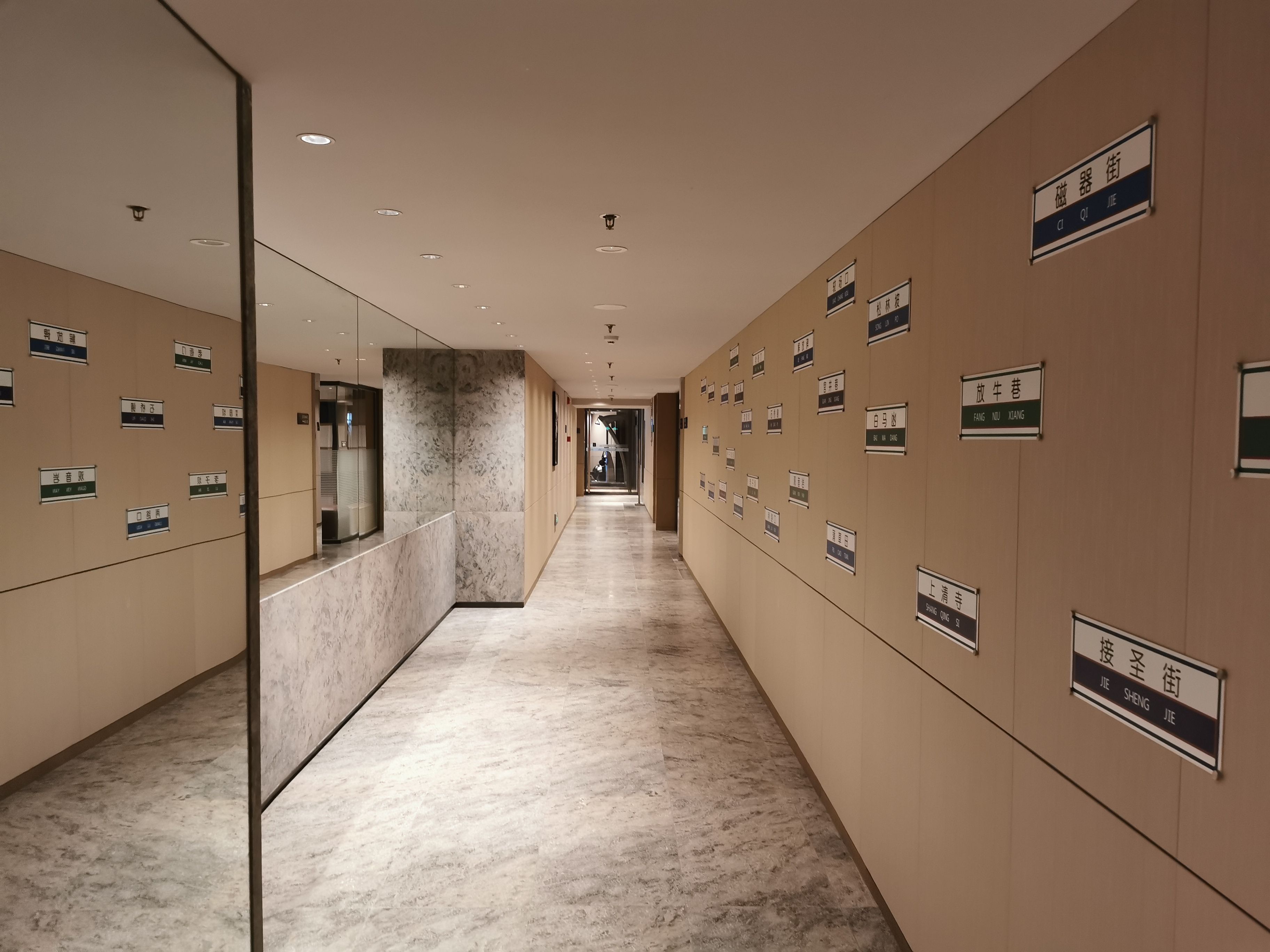 A hotel corridor with a marble floor, wall-mounted nameplates, and a glass elevator.