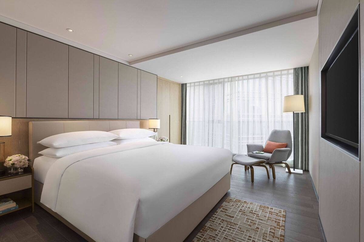 Ein modernes Hotelzimmer mit einem großen Bett, einer Sitzecke mit einem Sessel, einem Fernseher und einem großen Fenster mit Vorhängen.