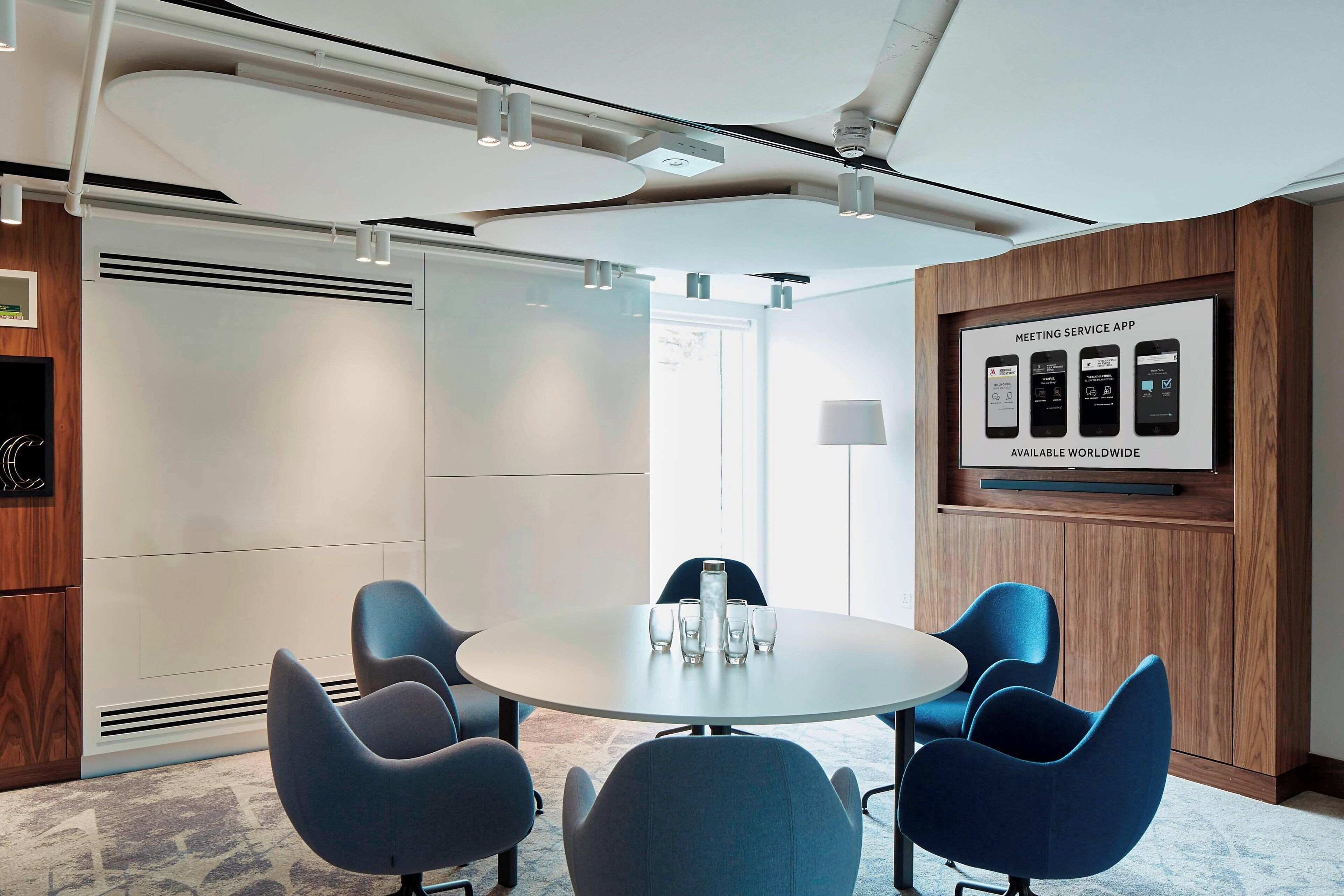 A modern meeting room with a round table, blue chairs, and a poster advertising a meeting service app.