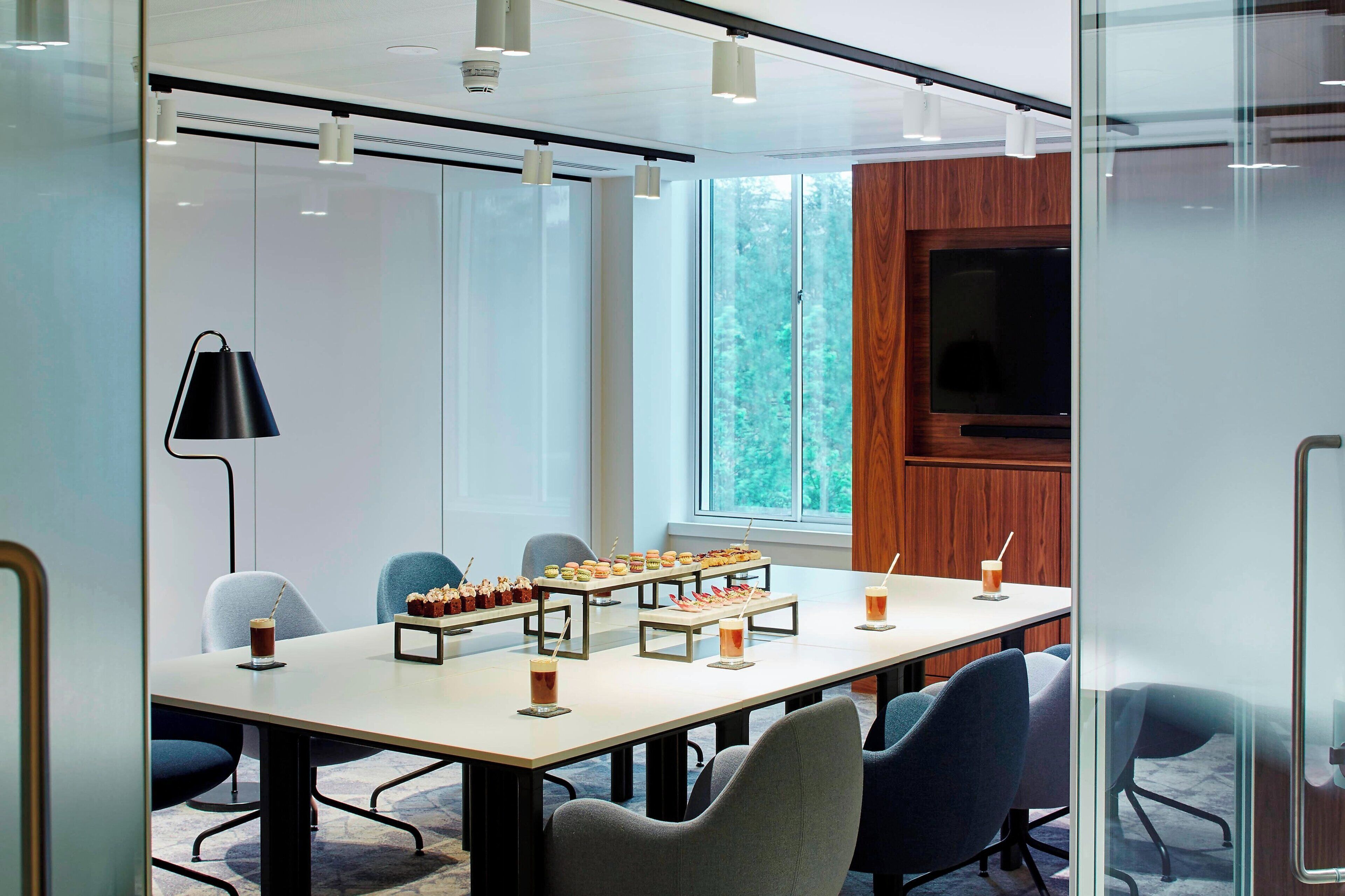 A conference room with a long table, chairs, a projector screen, and a window with a view of trees.