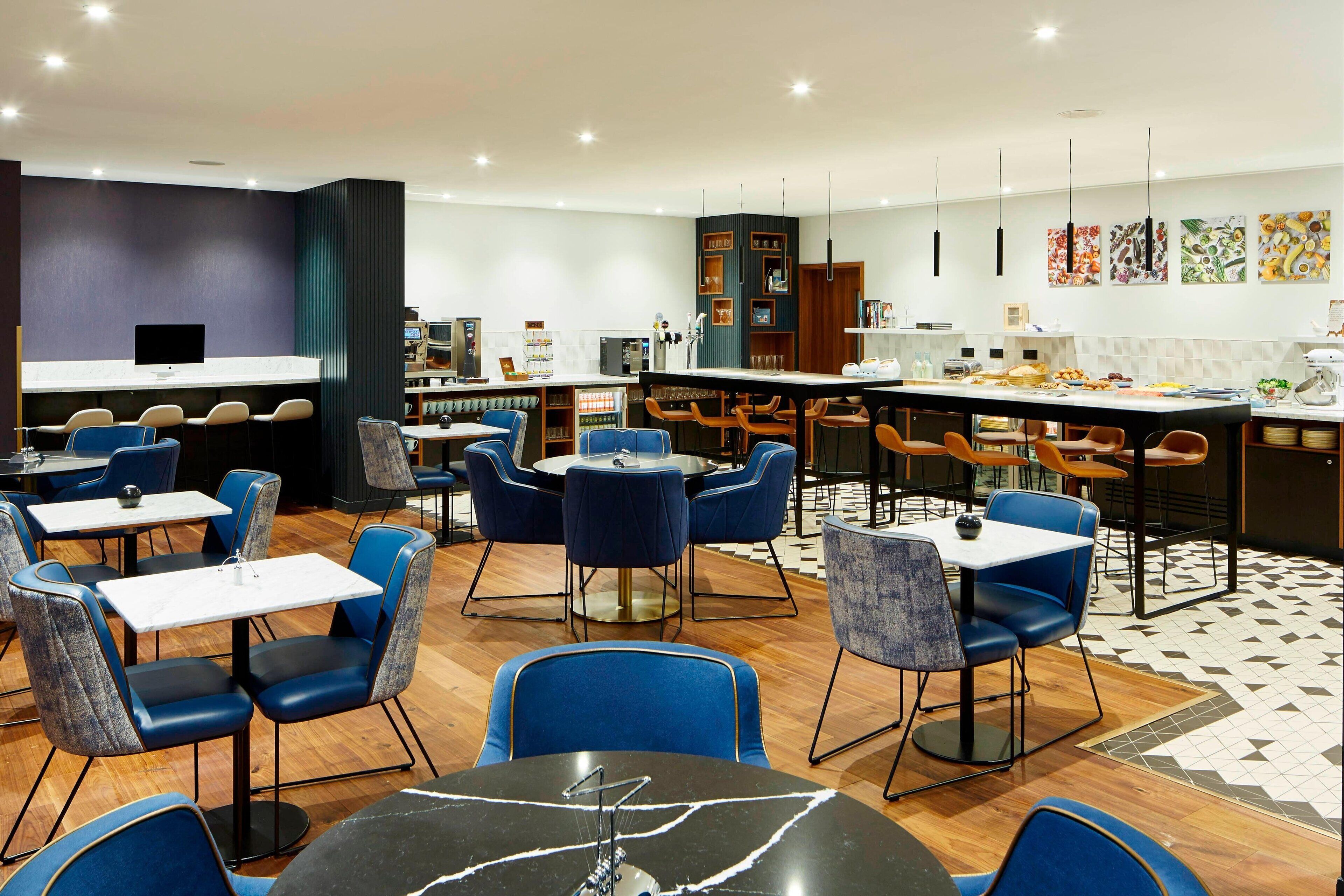 Een modern restaurant met blauwe en grijze stoelen, een bar in het midden en een zichtbare keuken op de achtergrond.