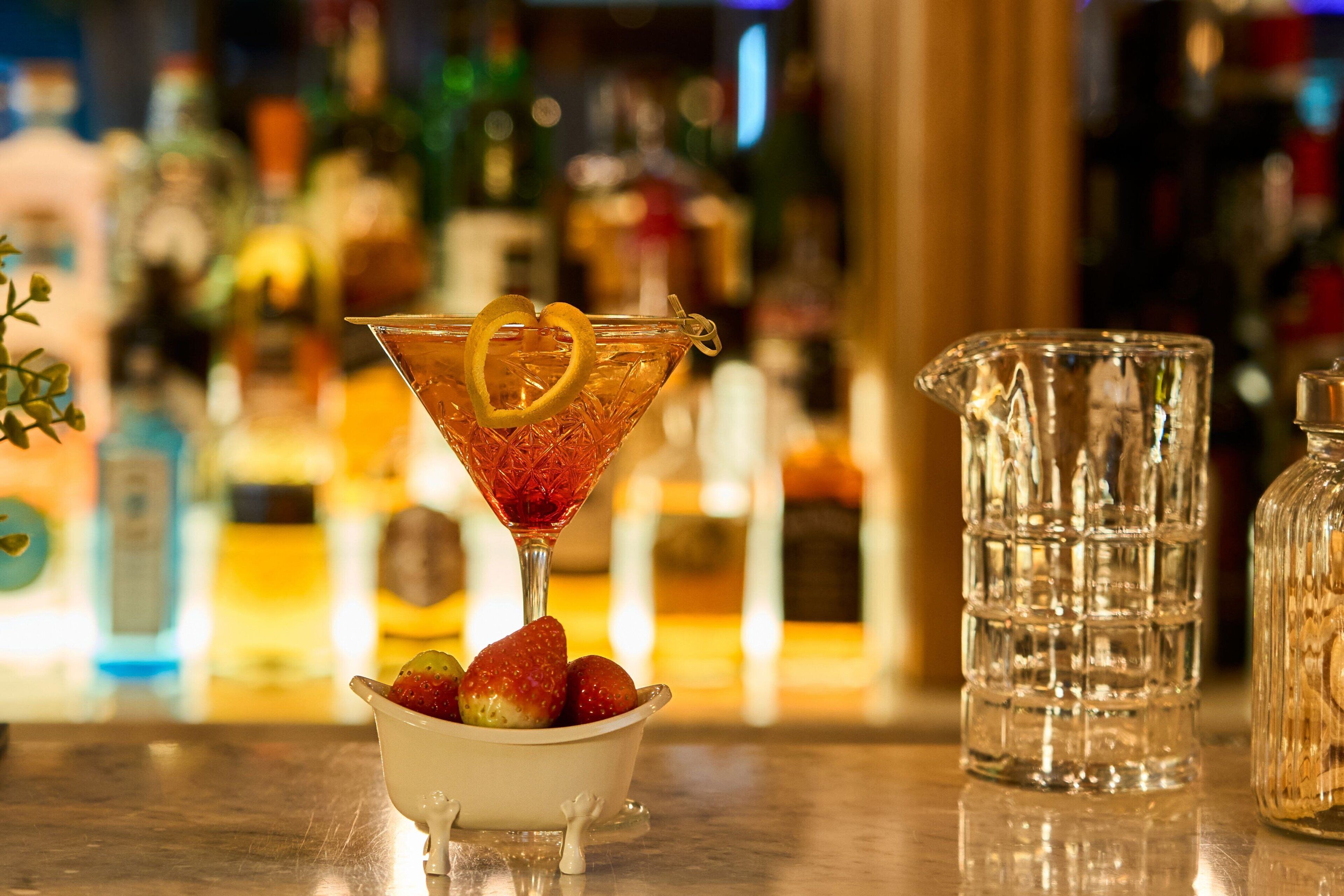 Ein Martini-Glas mit Zitronenscheibe und Erdbeeren auf einer Bar, im Hintergrund ein Krug und Flaschen.
