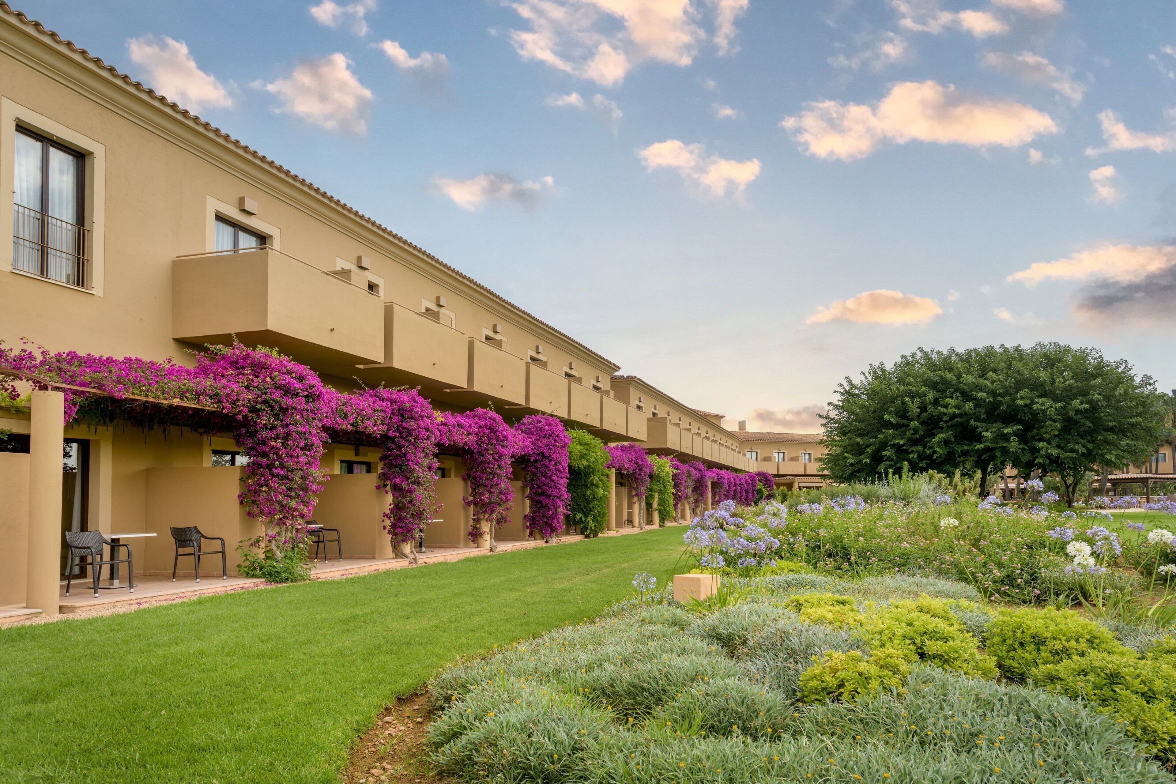 Un edificio con una larga hilera de balcones cubiertos de flores moradas, un césped bien cuidado y una variedad de plantas.