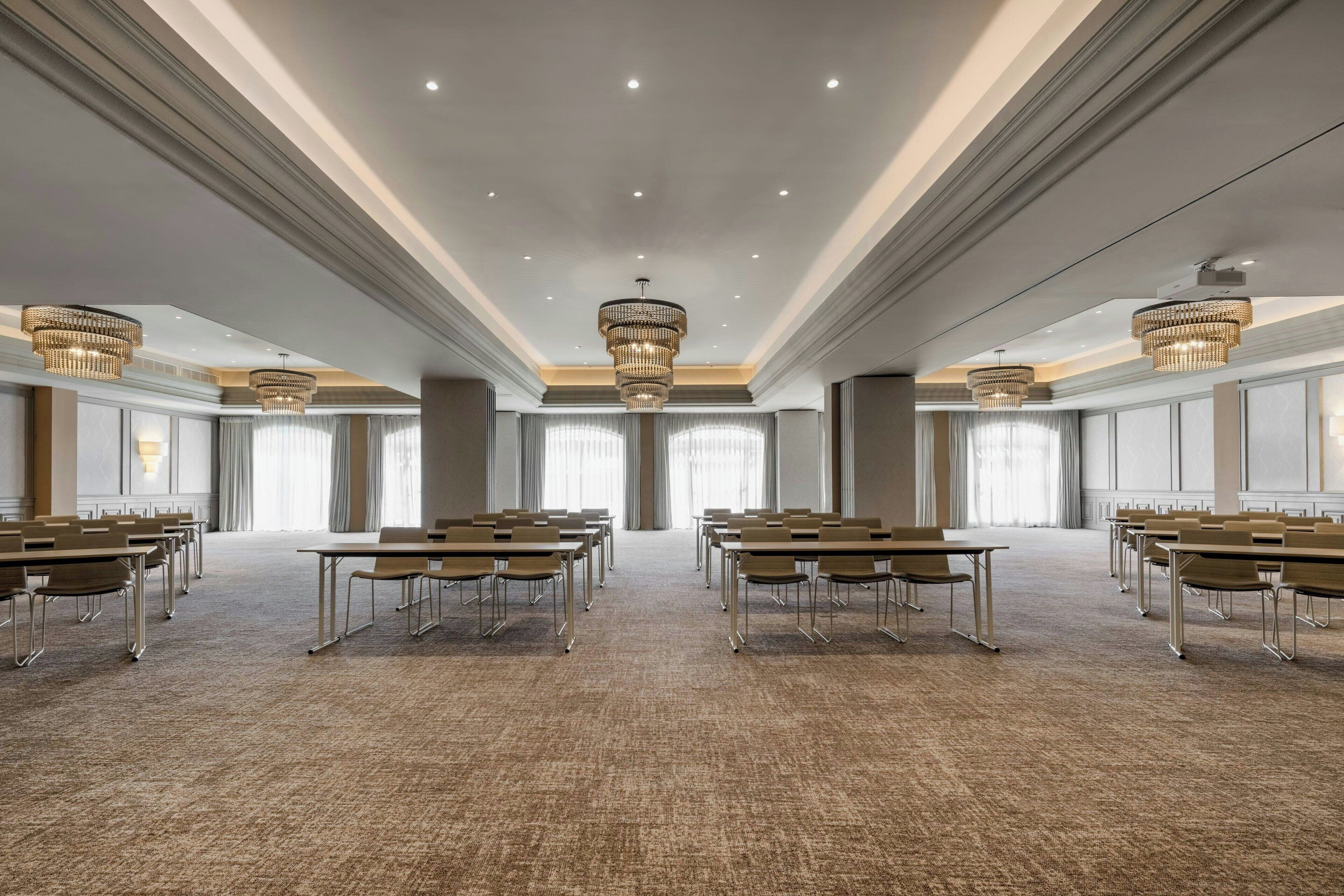 A conference room with rows of tables and chairs, large chandeliers, and a carpeted floor.