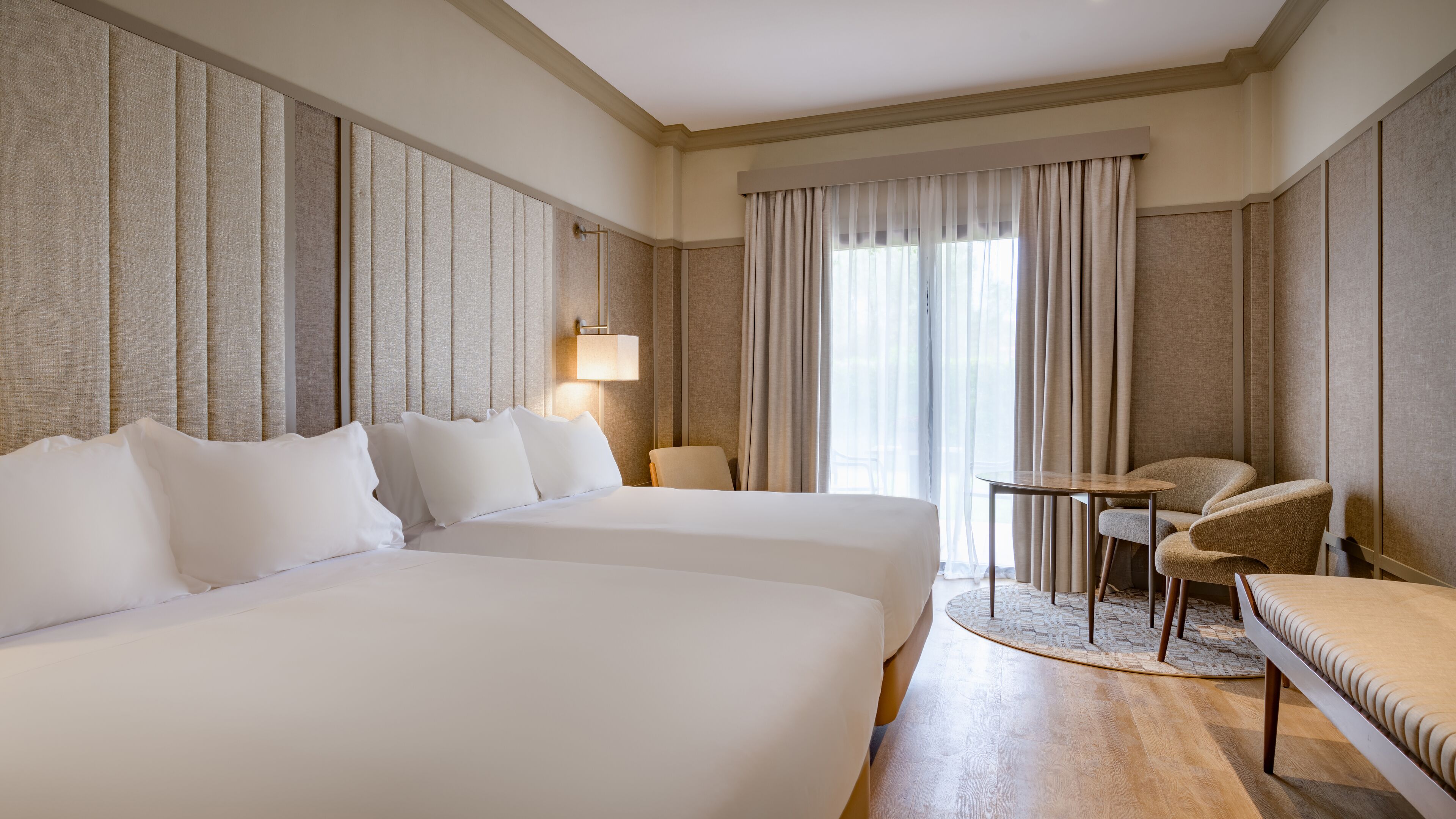 A hotel room with two beds, a small table, a chair, and a large window with curtains.