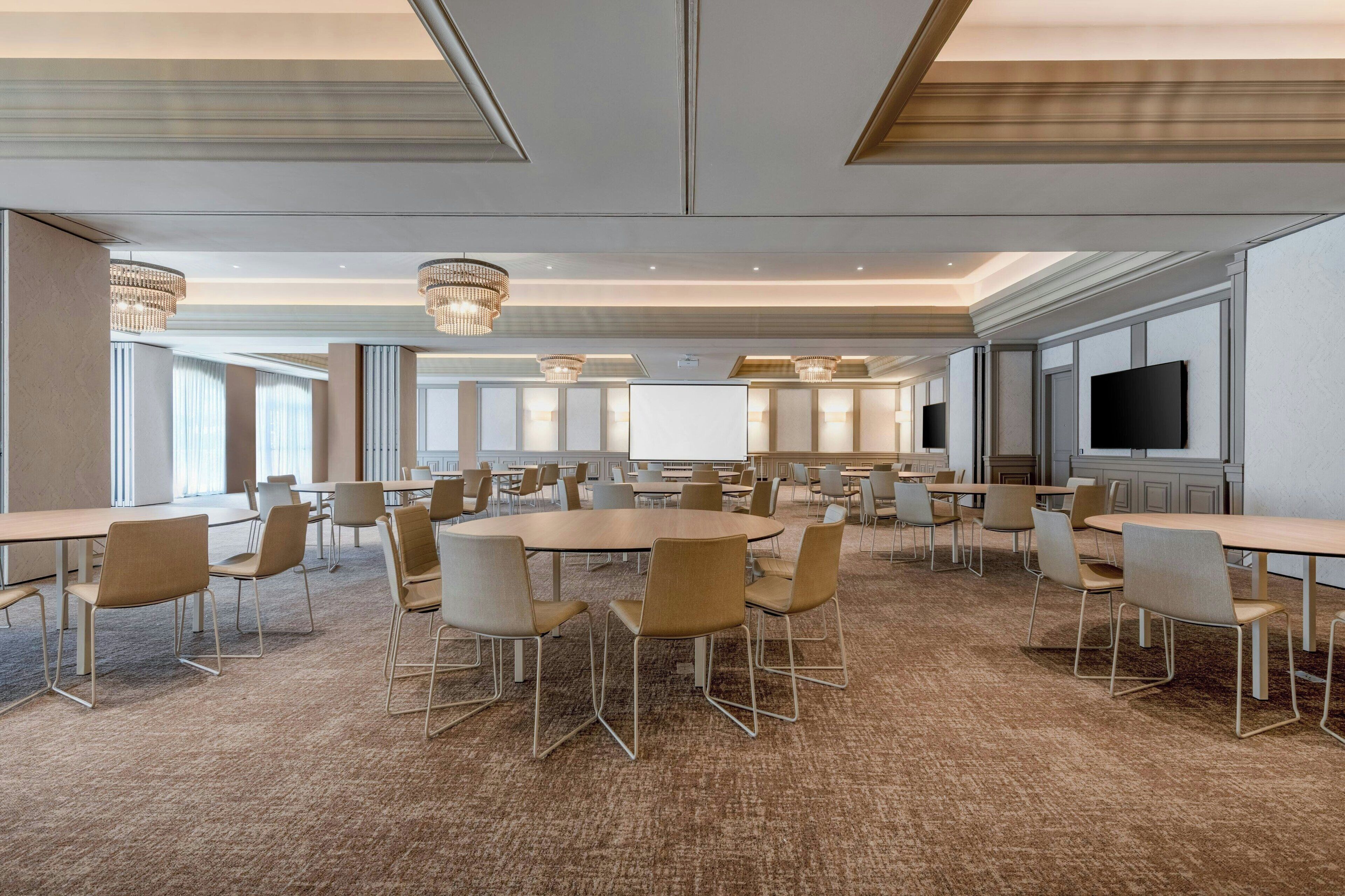 A spacious conference room with round tables and chairs, large windows, and a flat-screen TV.