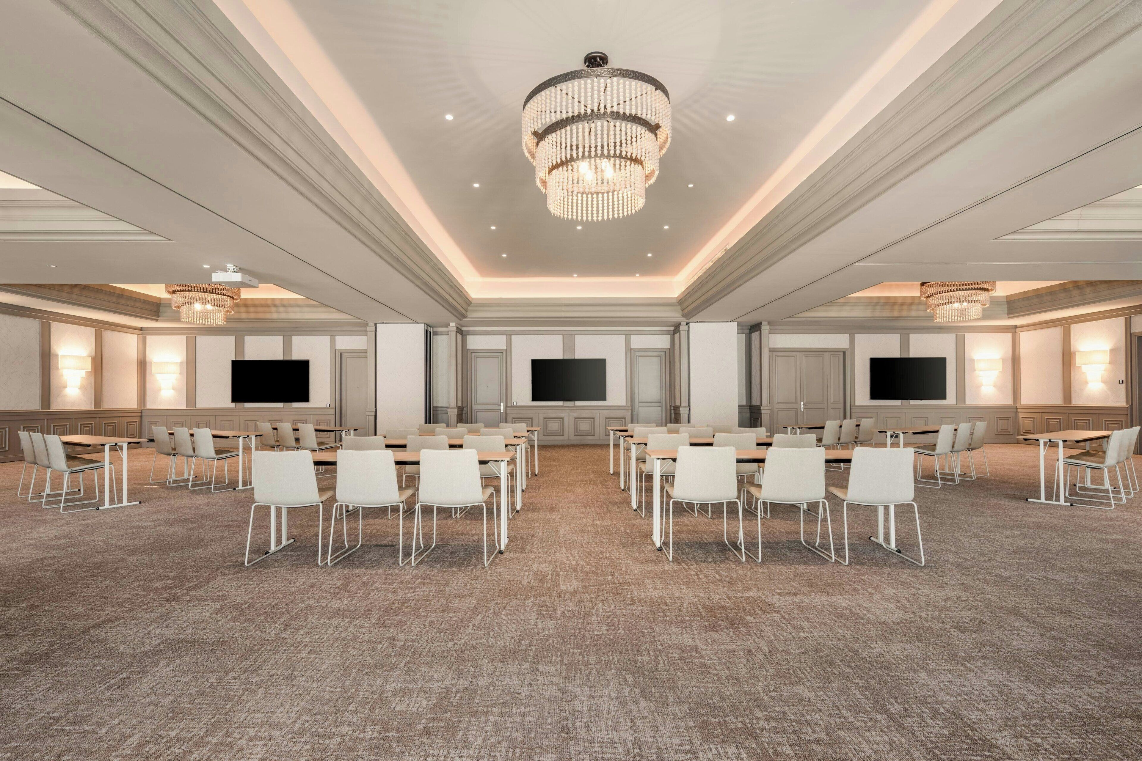 A conference room with a large chandelier, multiple tables with chairs, and two large flat-screen TVs on the walls.