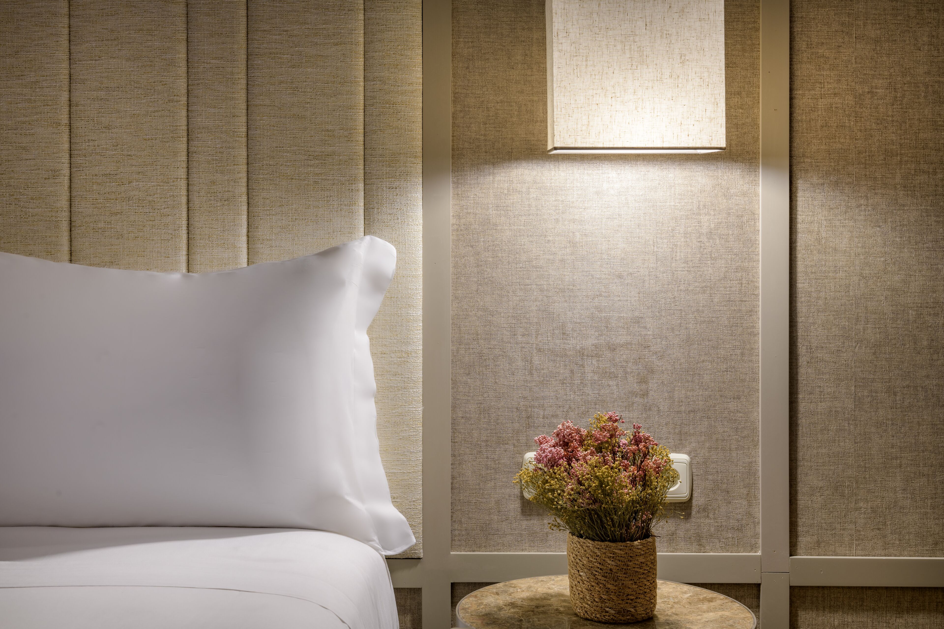 Una cama bien hecha con una almohada, una mesita de noche con una cesta tejida que contiene flores, y una lámpara de pared.