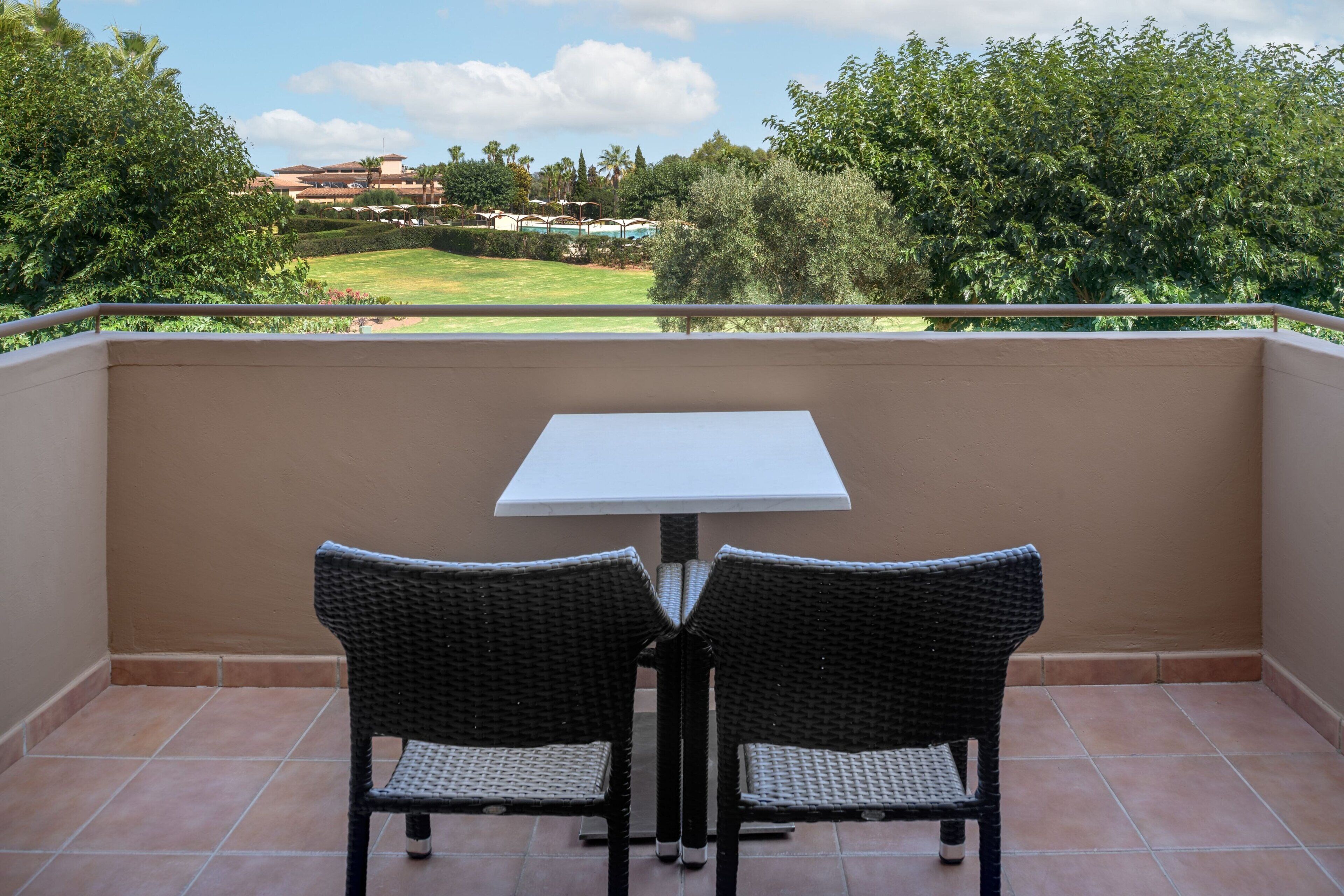 Un balcón con dos sillas de mimbre y una mesa blanca, con vistas a un jardín ajardinado y edificios a lo lejos.