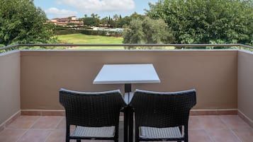 Un balcón con dos sillas de mimbre y una mesa blanca, con vistas a un jardín ajardinado y edificios a lo lejos.
