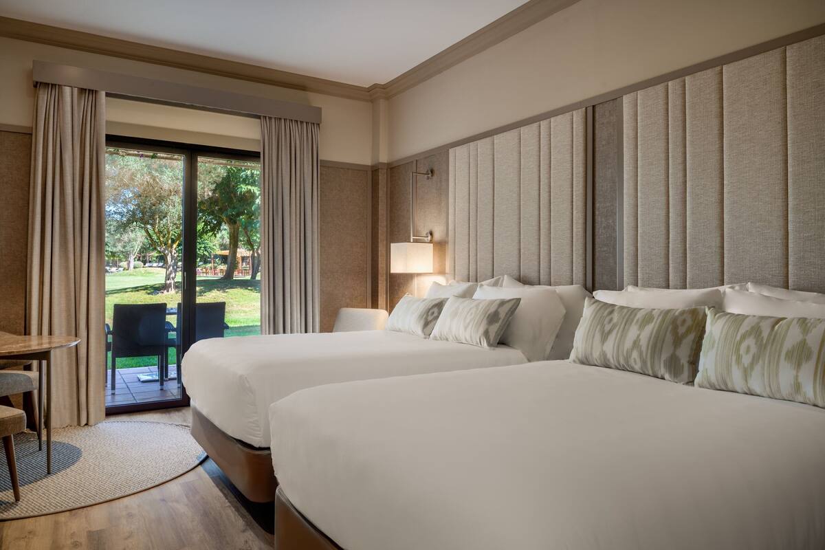 A hotel room with two beds, a desk, and a view of a garden.