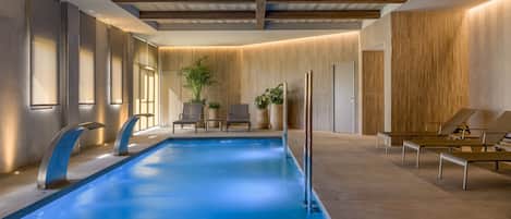 A modern indoor swimming pool with a clear blue water surface, wooden ceiling beams, and lounge chairs along the poolside.