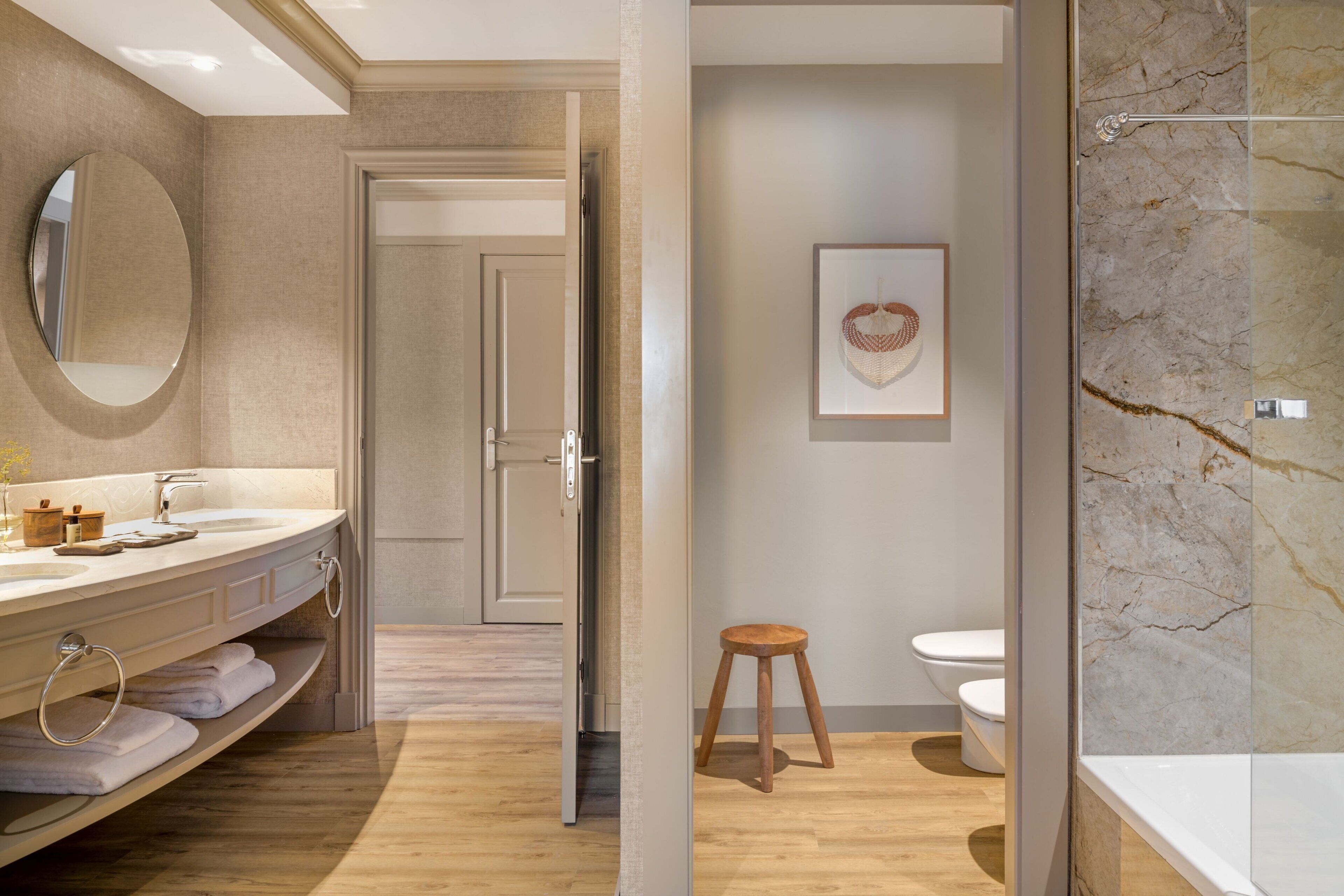 Un baño moderno con tocador de madera, un espejo redondo, una obra de arte enmarco y pared de ducha de mármol.