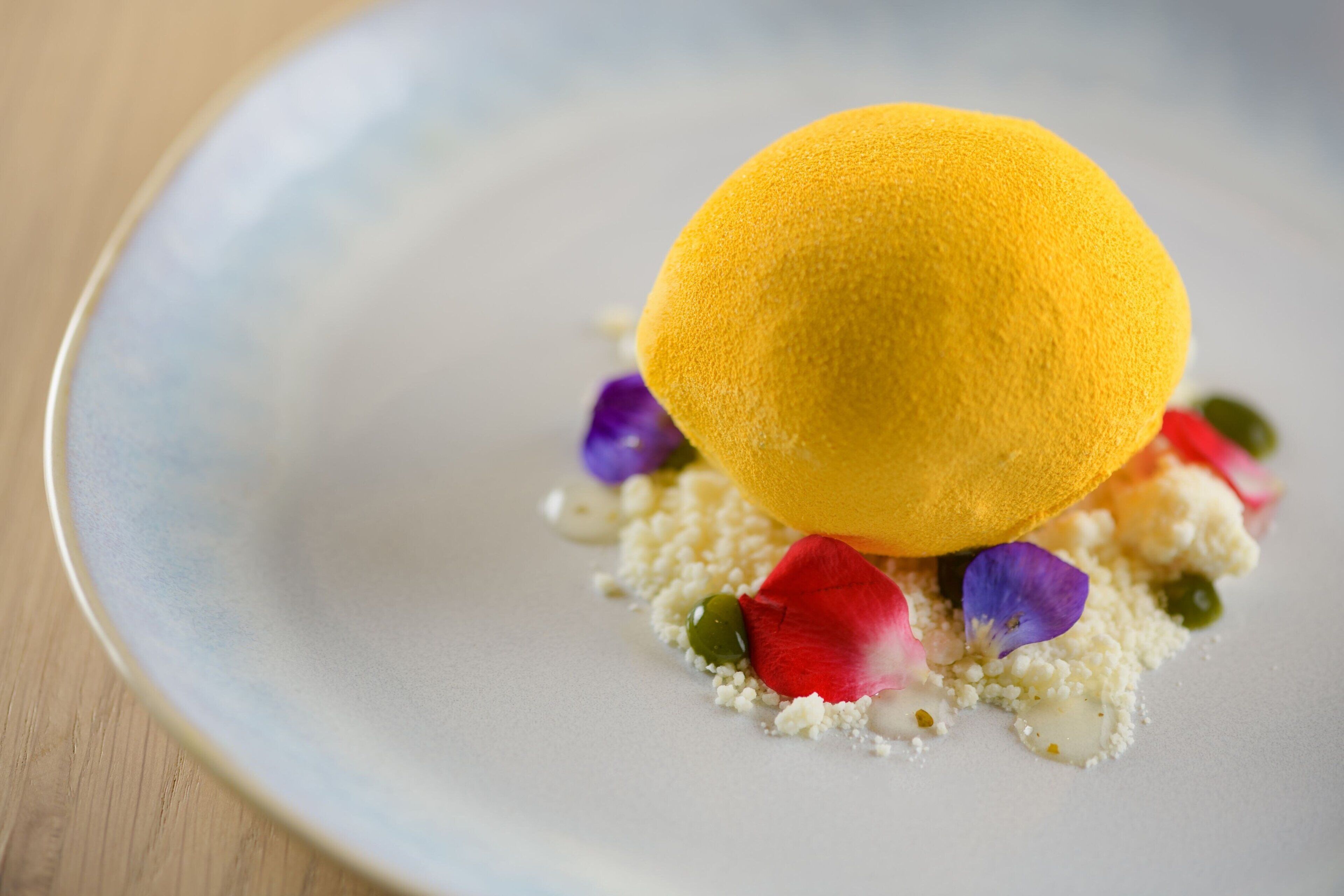 Un dessert composé d’une sphère jaune, présentée sur une assiette parsemée d’éléments blancs et verts, ainsi que de pétales de fleurs violettes.
