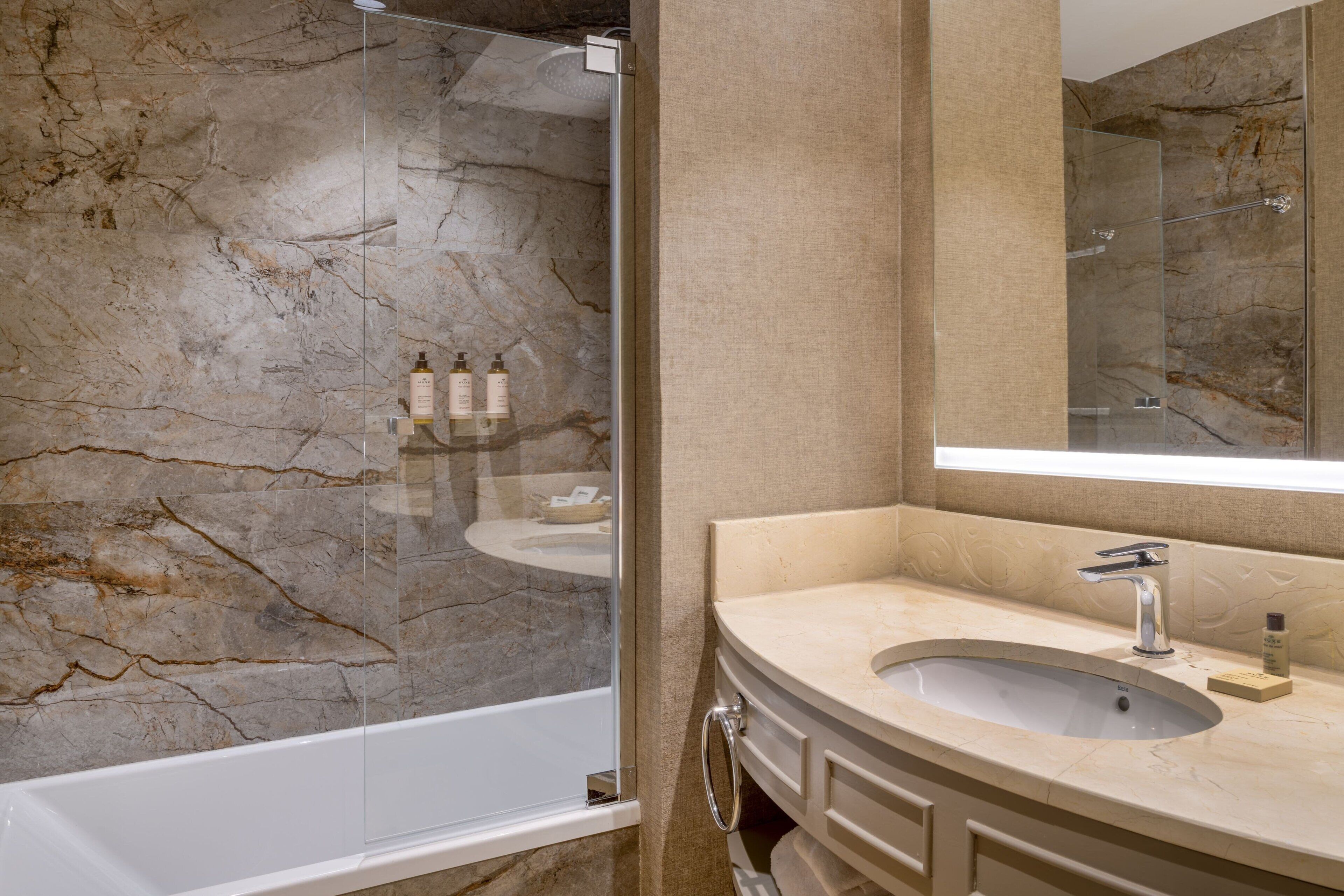 Un baño moderno con pared de mármol, ducha con cerramiento de vidrio, lavabo con espejo y tocador con armarios.
