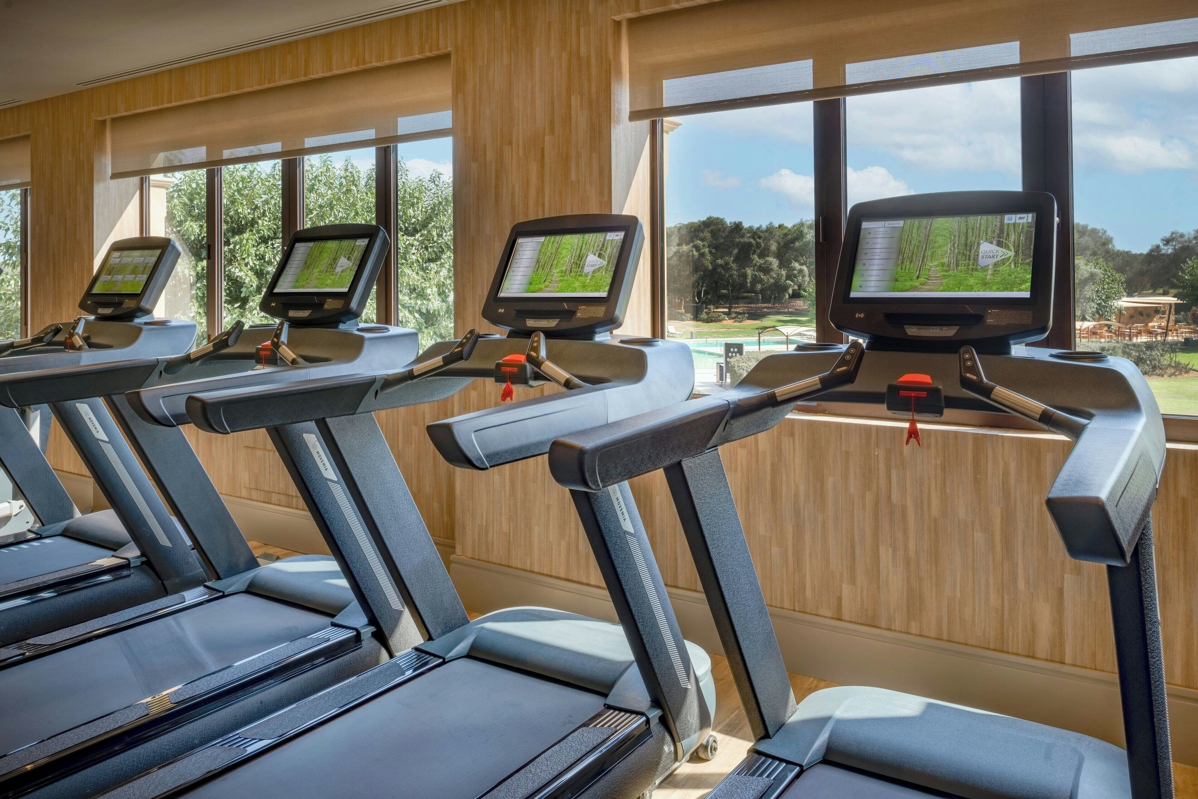 Ein Fitnessraum mit Laufbändern, jeweils ausgestattet mit einem digitalen Bildschirm, großen Fenstern und Holzwänden.