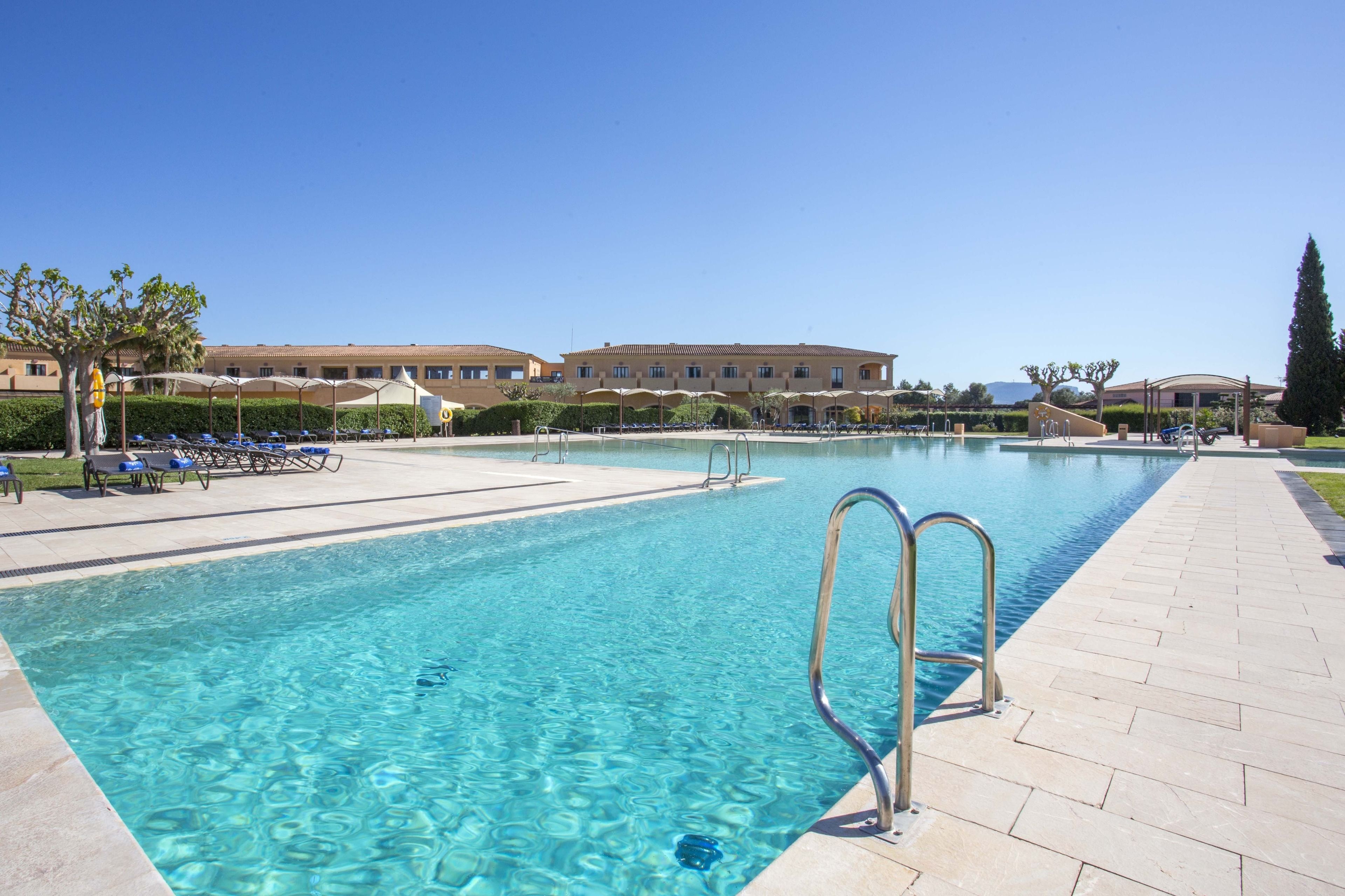 A clear swimming pool with lounge chairs and umbrellas, surrounded by a landscaped area and a building in the background.