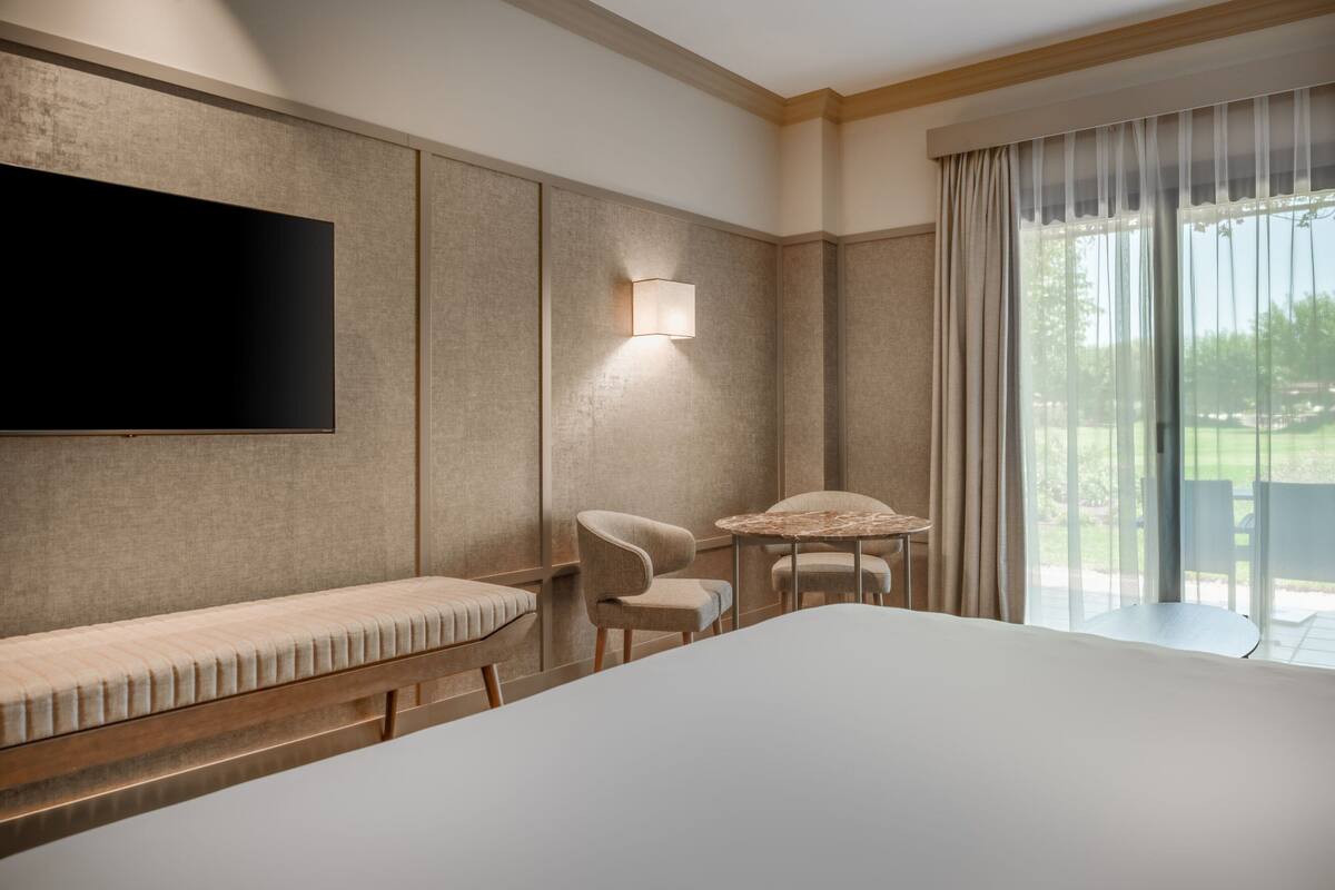 A modern hotel room with a large bed, a flat-screen TV mounted on the wall, a small table with chairs, and a large window with curtains.