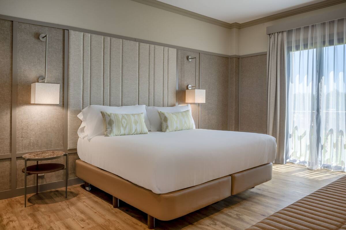 A modern hotel room with a large bed, two bedside lamps, a small table, and a view through sheer curtains.