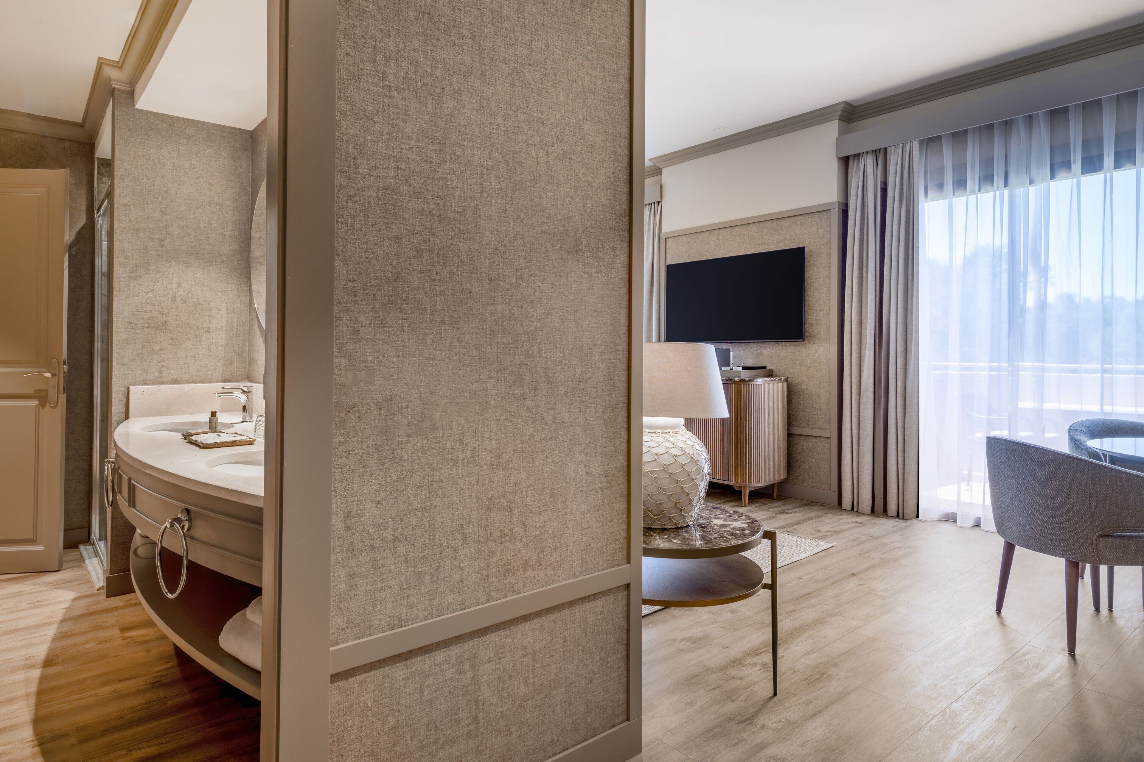 Una habitación de hotel moderna con una cama grande, un área de descanso con una mesa y sillas, un televisor montado en la pared y un baño con lavamanos y espejo.