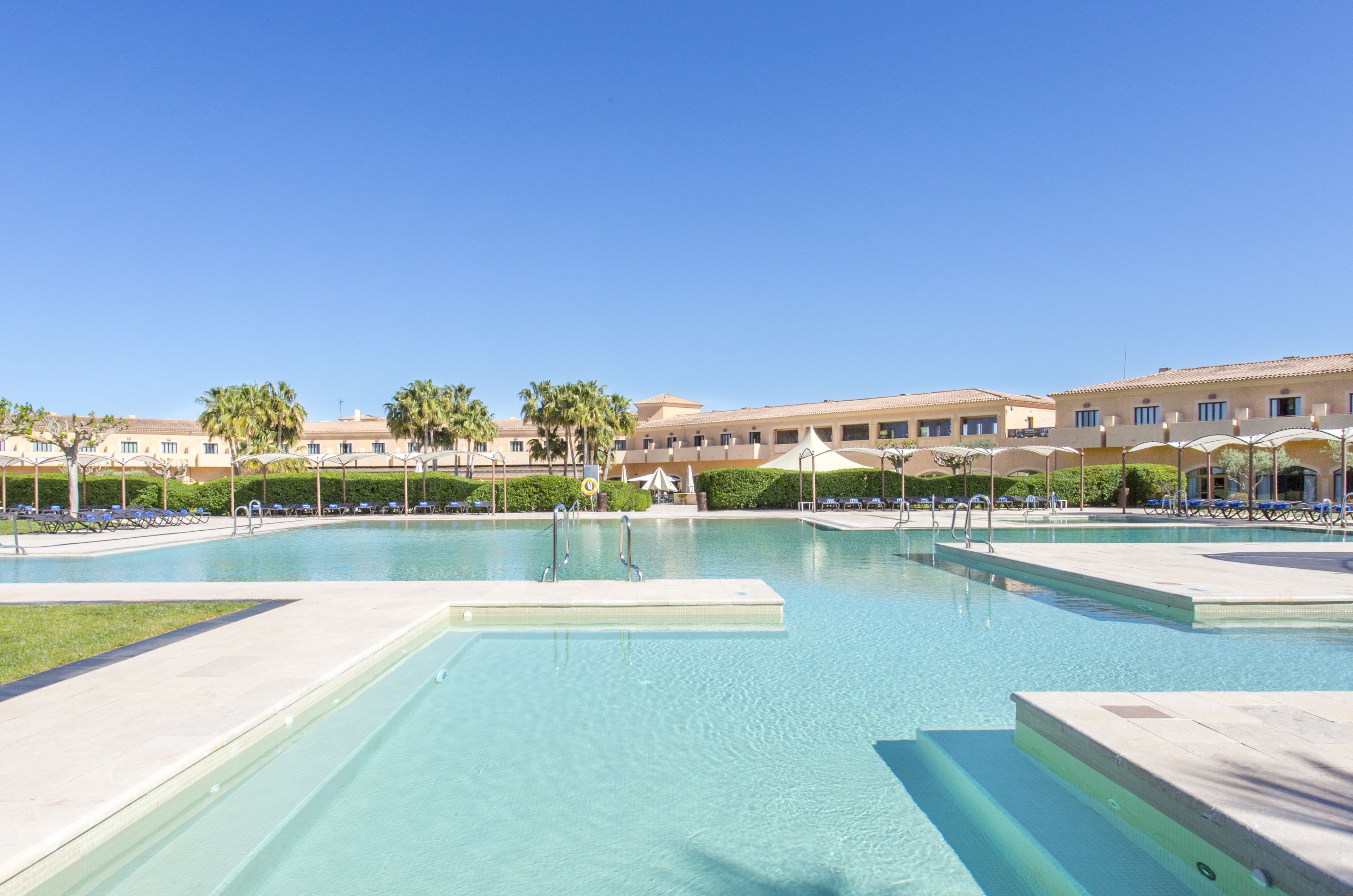 A swimming pool with clear blue water, surrounded by lounge chairs and umbrellas, and a building with multiple balconies in the background.