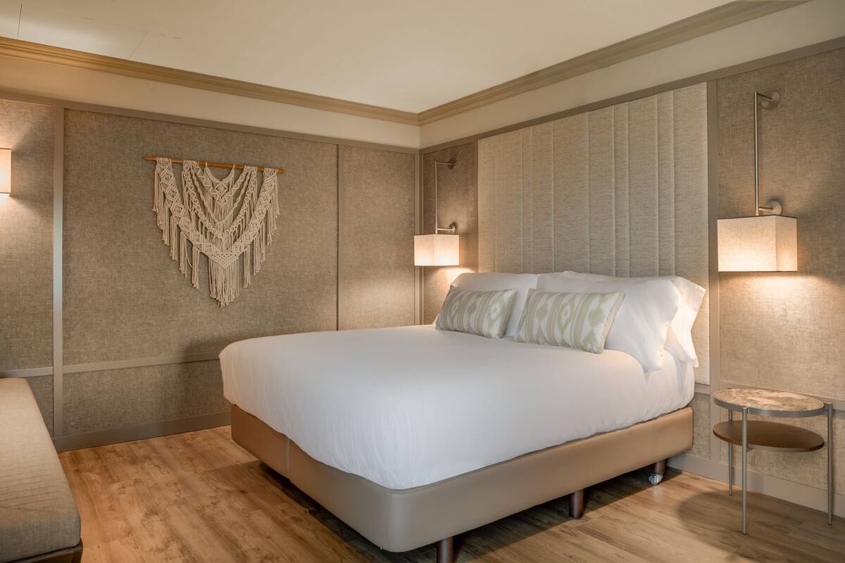 A modern hotel room with a large bed, a bedside table, a wall-mounted macrame piece, and two wall-mounted lamps.