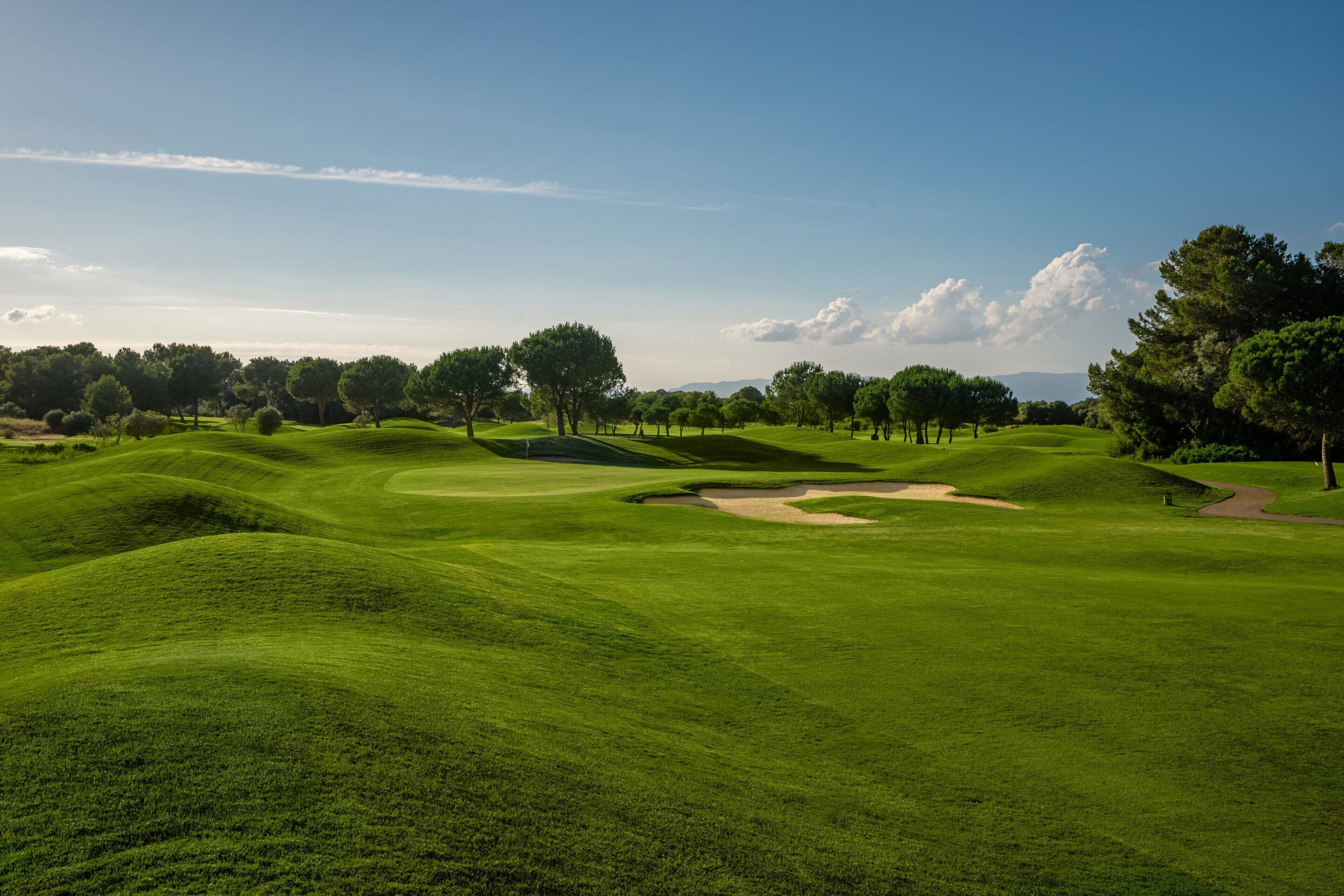 A golf course with rolling green hills, sand bunkers, and trees under a clear sky.