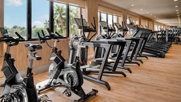 Uma academia com diversas máquinas de cardio, janelas amplas e piso de madeira.