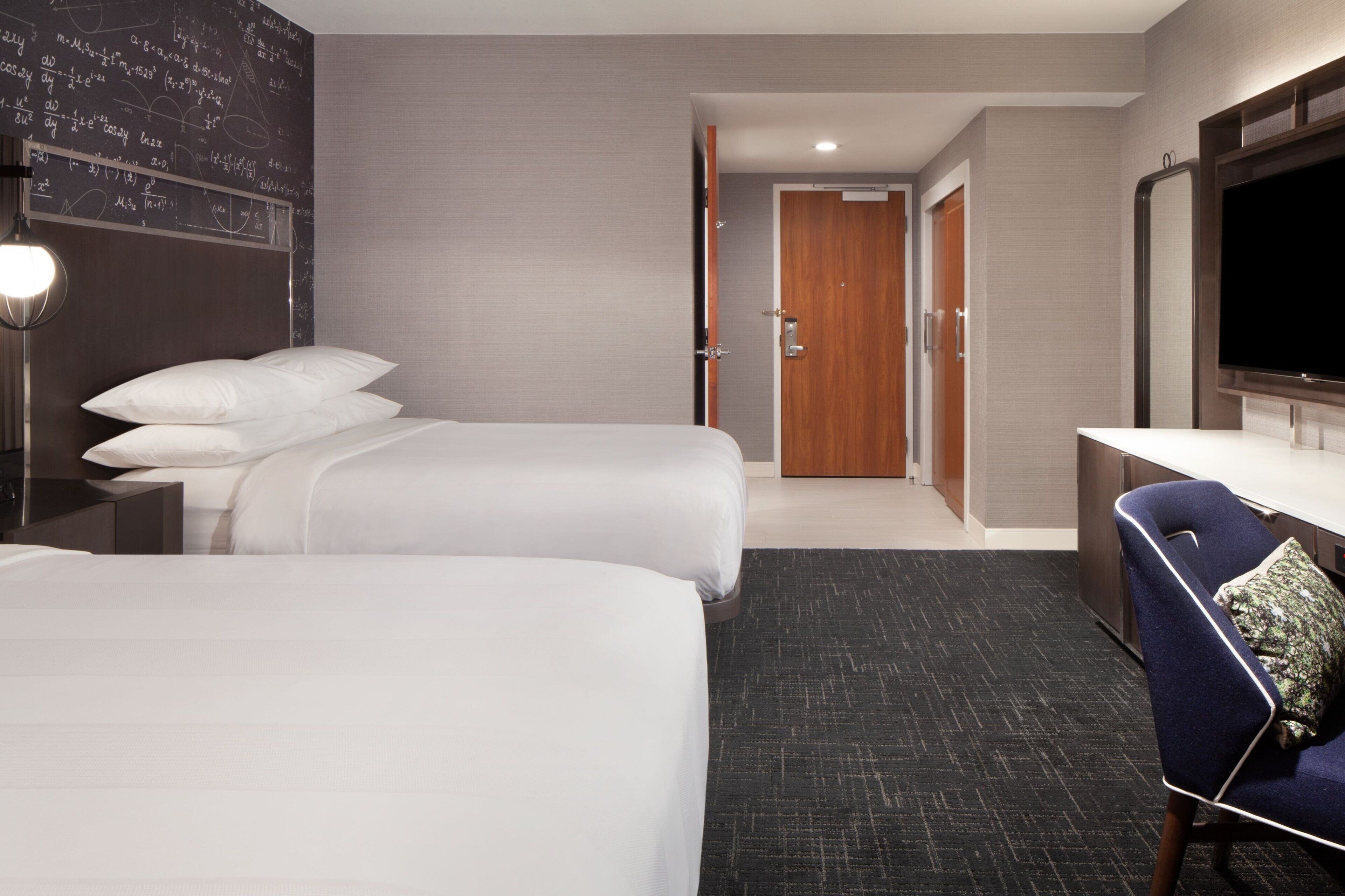 A hotel room with two beds, a desk, a chair, a TV, and a door leading to a bathroom.