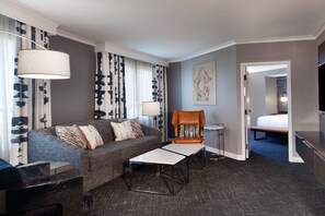 A hotel room with a grey sofa, a brown armchair, a small white table, a lamp, and a bed in the adjacent room. - Fremont Marriott Silicon Valley (Fremont)