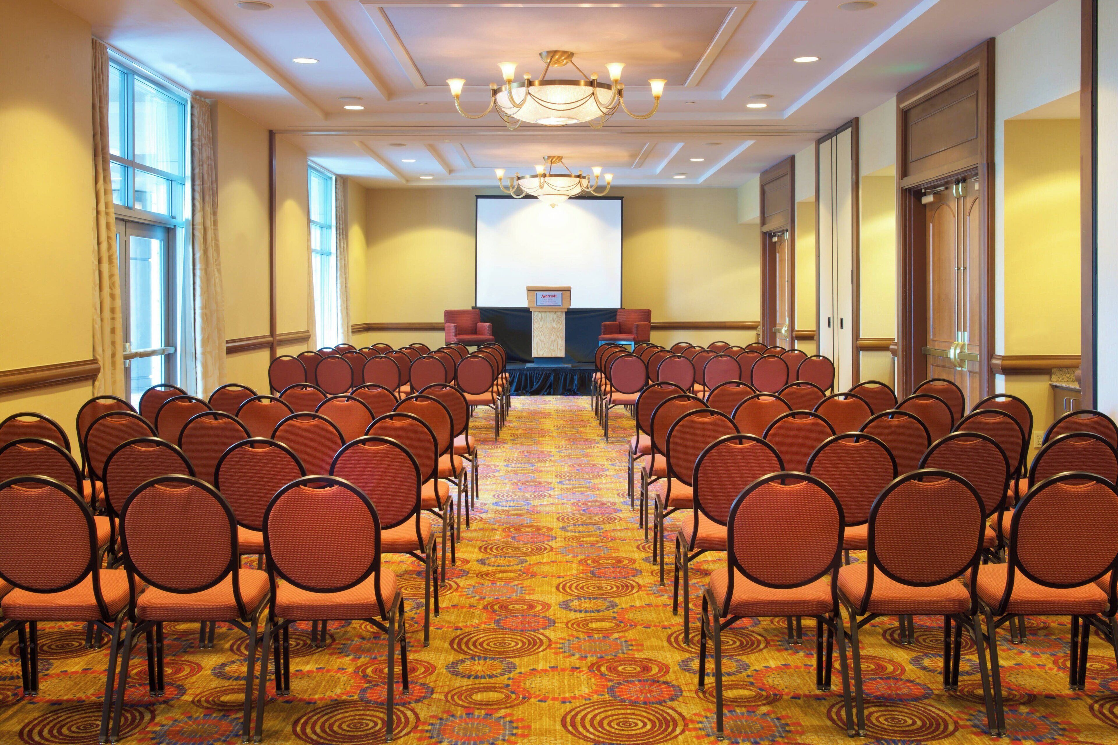 A conference room with rows of chairs facing a podium, large windows, and a chandelier.