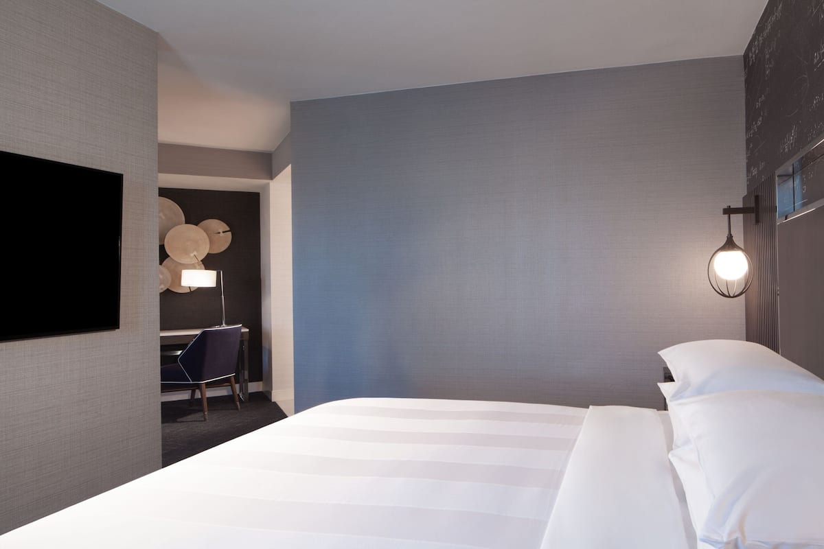 Habitación de hotel con una cama grande, un televisor de pantalla plana en la pared, un escritorio con una silla y una lámpara de pared.
