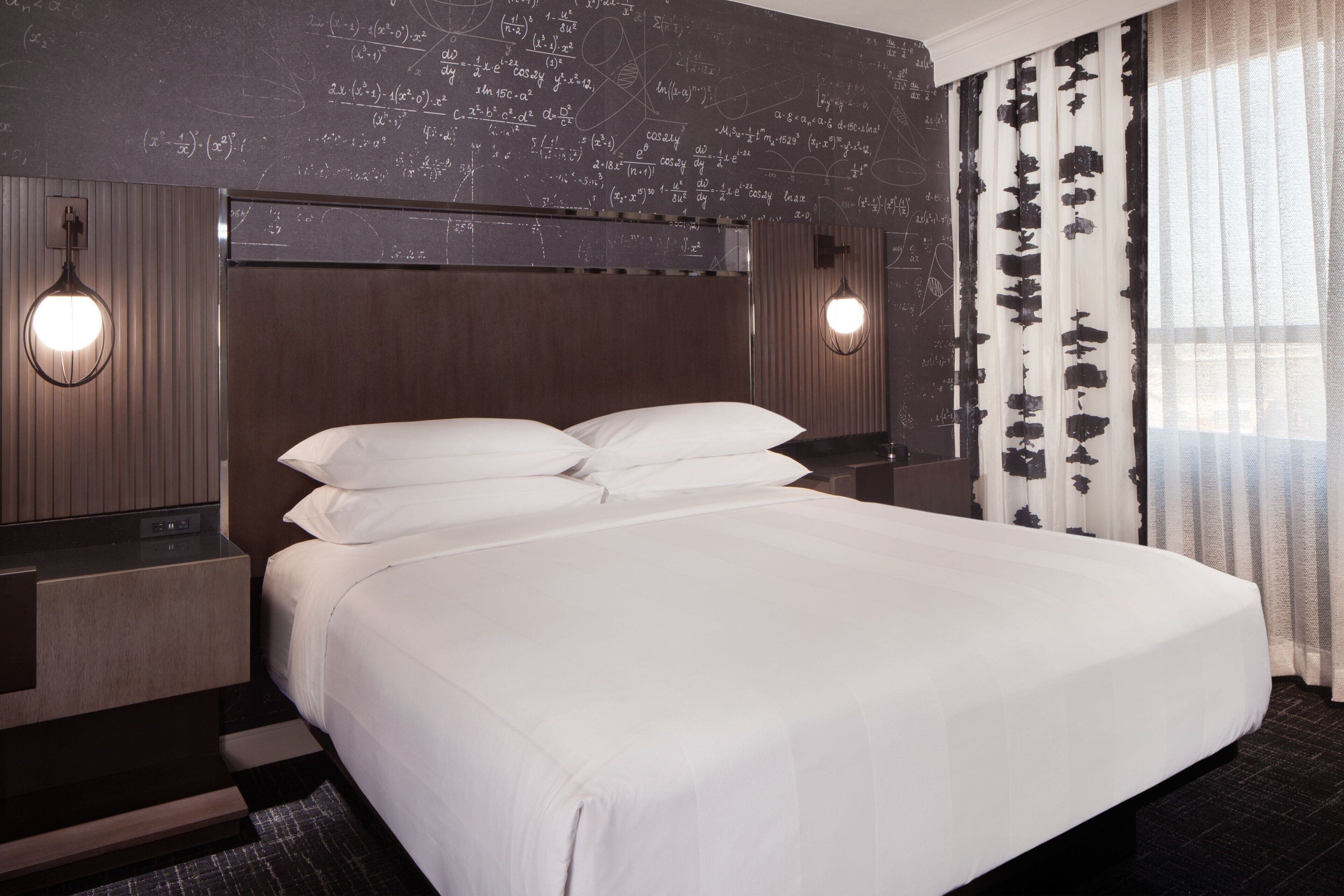 Ein Hotelzimmer mit einem großen Bett, zwei Nachttischen, einem Kopfteil mit einer Leuchte und einer Wand mit mathematischen Gleichungen.