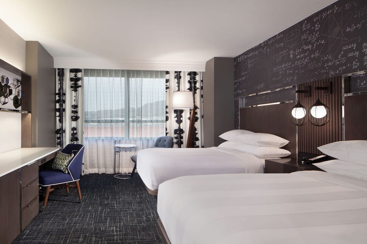Habitación de hotel con dos camas, un escritorio, una silla, una lámpara y vista al exterior.