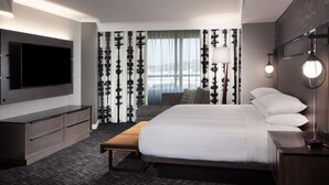 Une chambre d’hôtel moderne dotée d’un grand lit, d’un téléviseur à écran plat fixé au mur, d’une table de chevet avec une lampe et d’une fenêtre ornée de rideaux légers.