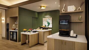 A modern kitchen area with a coffee machine, fruit bowls, and a refrigerator.