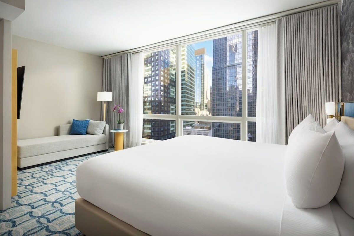 A hotel room with a large bed, a sofa, a lamp, and a view of tall buildings.