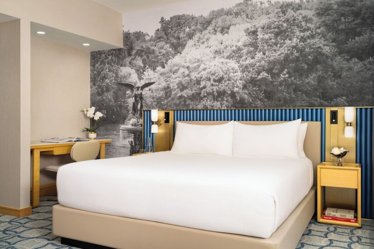 A hotel room with a bed, a desk, a chair, and a nightstand with a lamp.