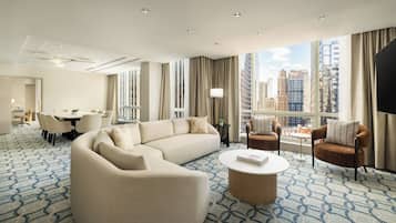 A modern hotel room with a large sectional sofa, a round coffee table, and a dining area with chairs.