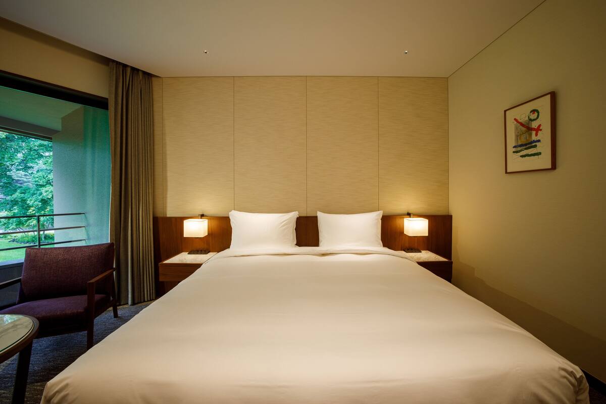 Habitación de hotel con una cama grande, dos mesitas de noche con lámparas, una silla y vistas a zonas verdes.