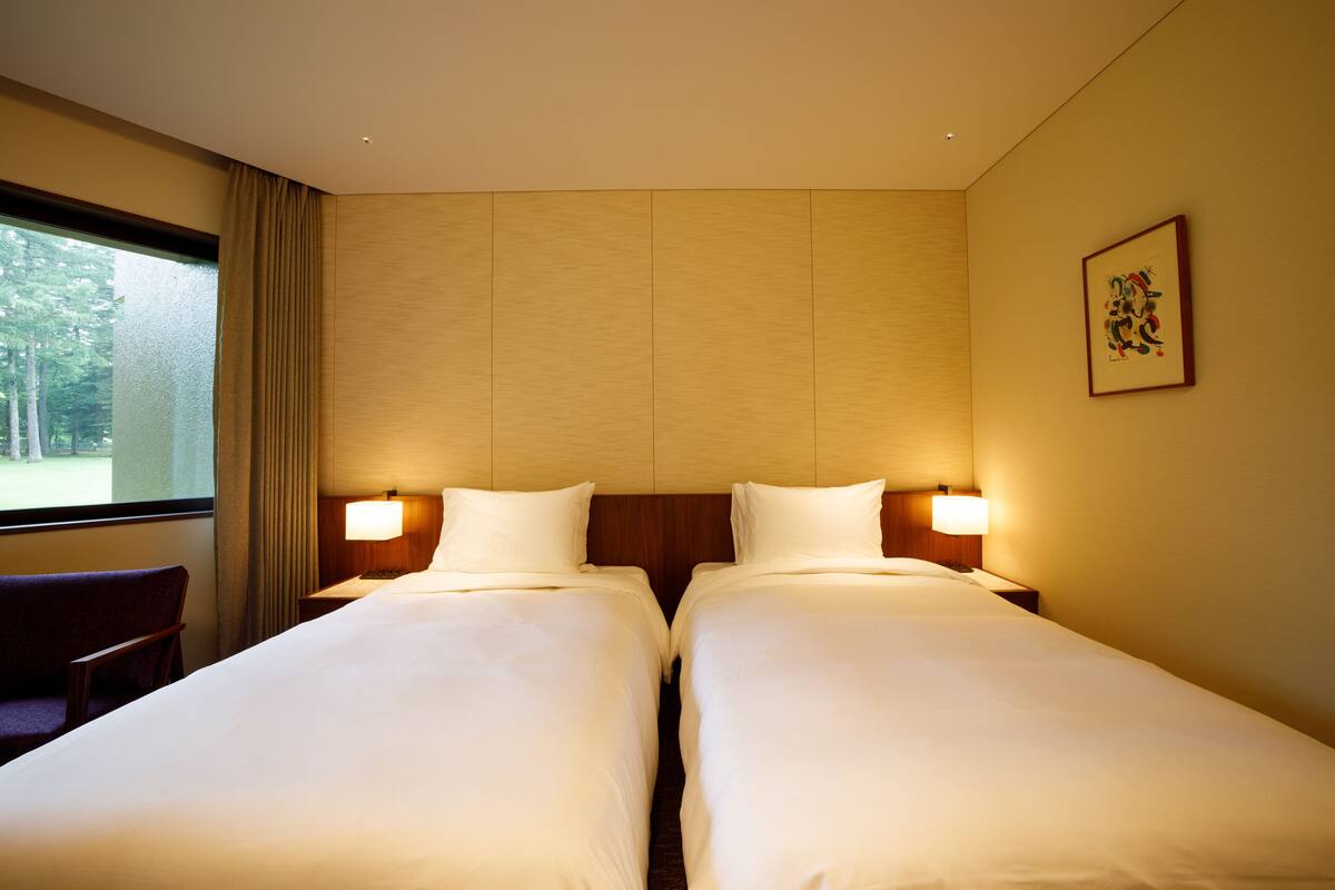 Habitación de hotel con dos camas, una ventana con cortinas, una silla y un cuadro en la pared.