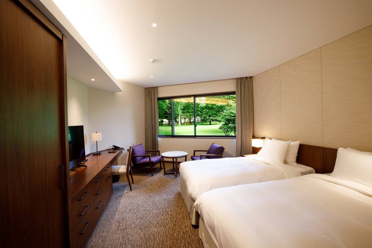 Habitación de hotel con dos camas, un escritorio con silla, un televisor y ventana con vistas a zonas verdes.
