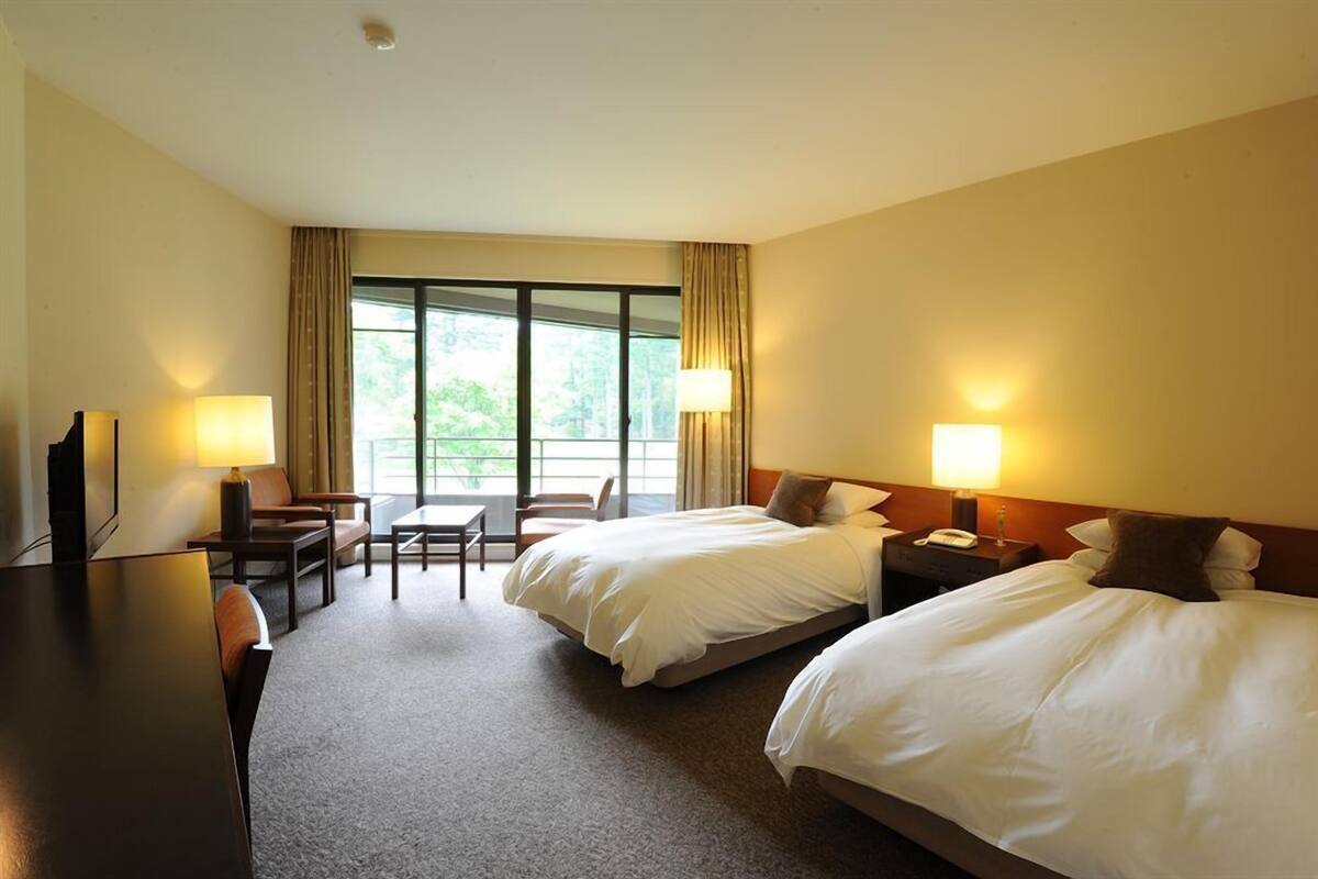 Habitación de hotel con dos camas, un escritorio, una silla y un ventanal con cortinas.