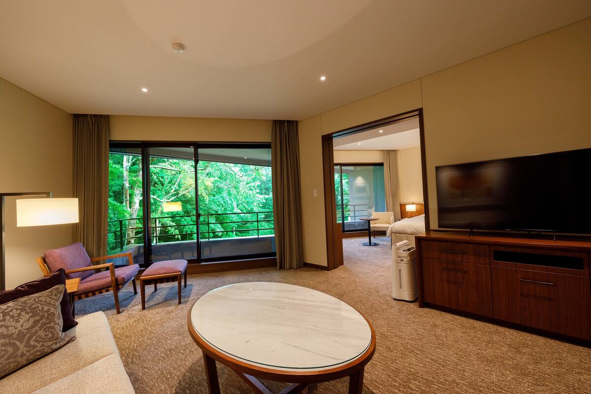 Habitación de hotel moderna con un televisor grande, asientos cómodos, un balcón con vistas y suelo alfombrado.