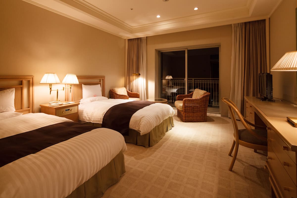 A hotel room with two beds, a desk, a chair, a TV, and a balcony with a table and chairs.