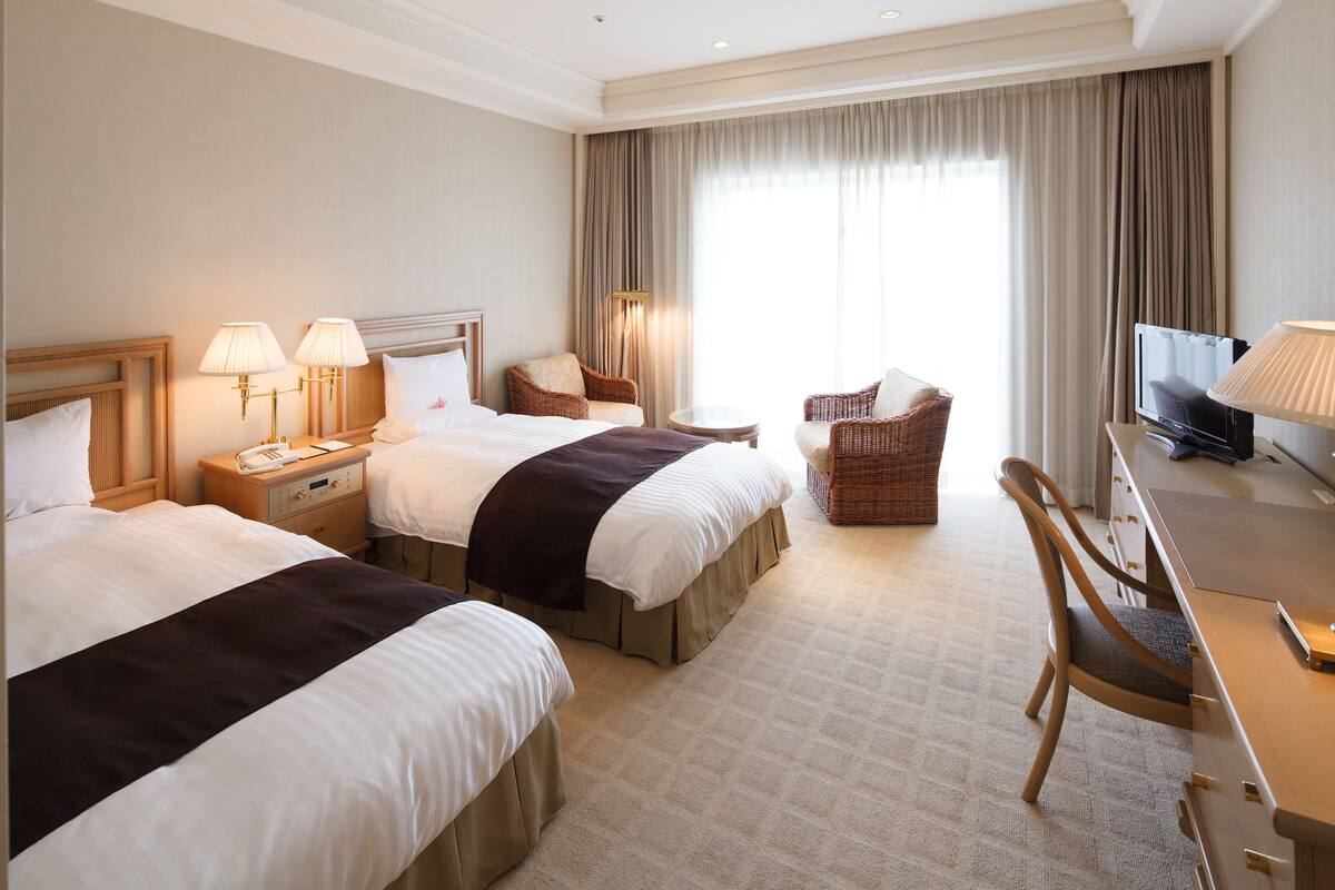 A hotel room with two beds, a desk, a chair, a television, and a window with curtains.