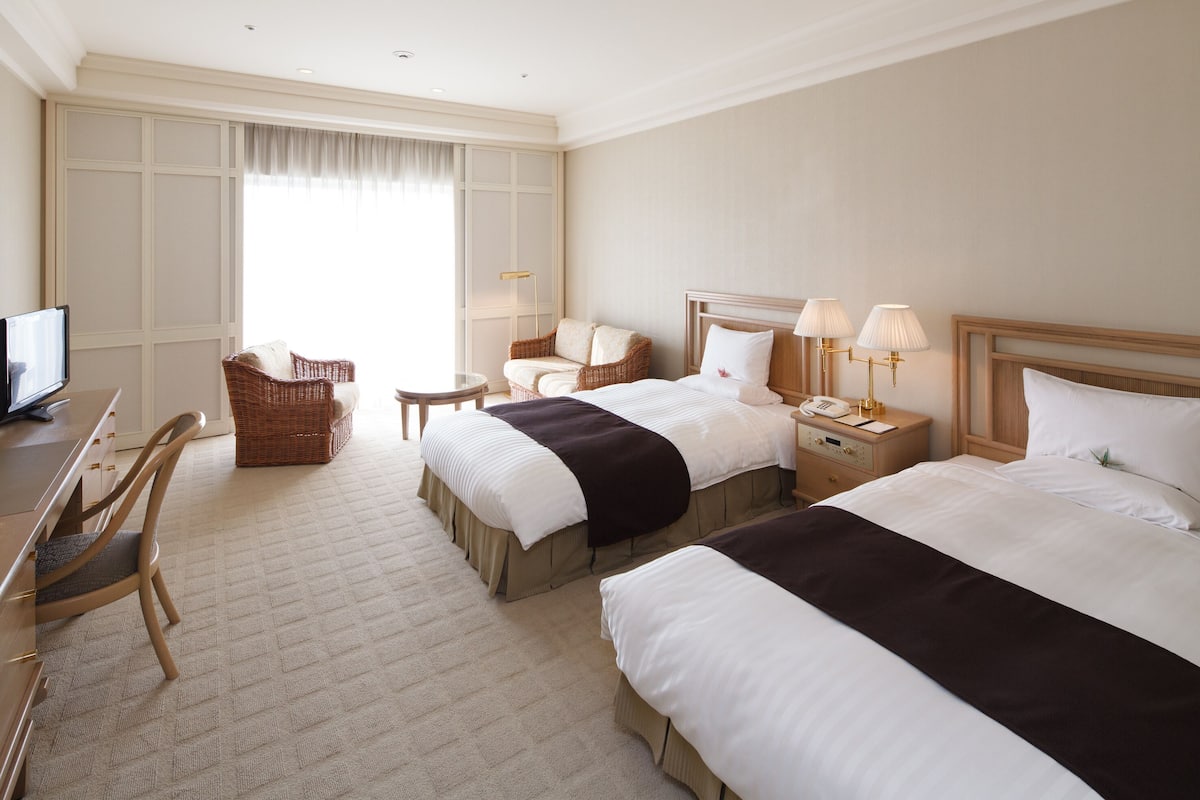A hotel room with two beds, a desk, a chair, a TV, and a window with curtains.