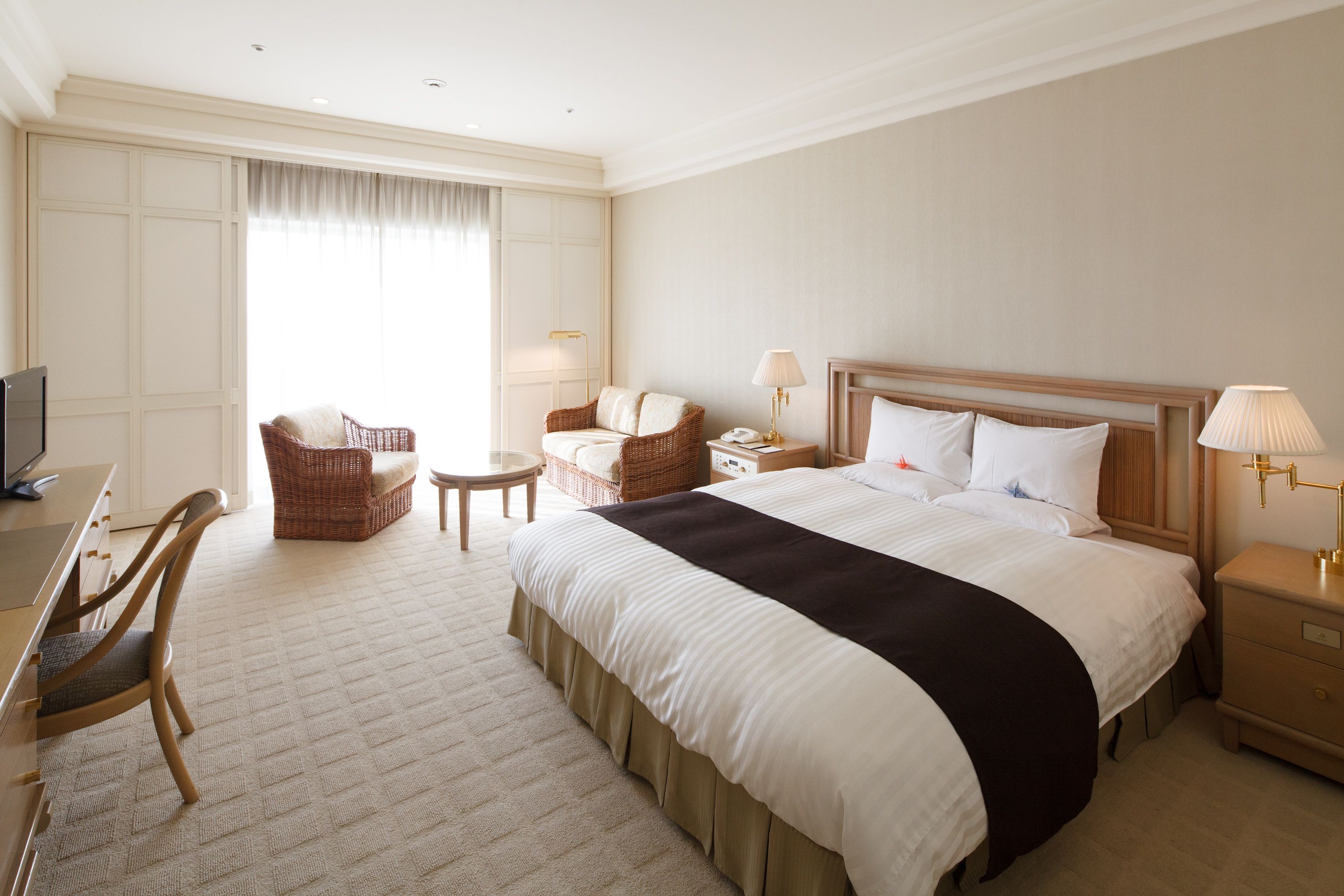 A hotel room with a large bed, a desk with a chair, a small round table, and a wicker chair.